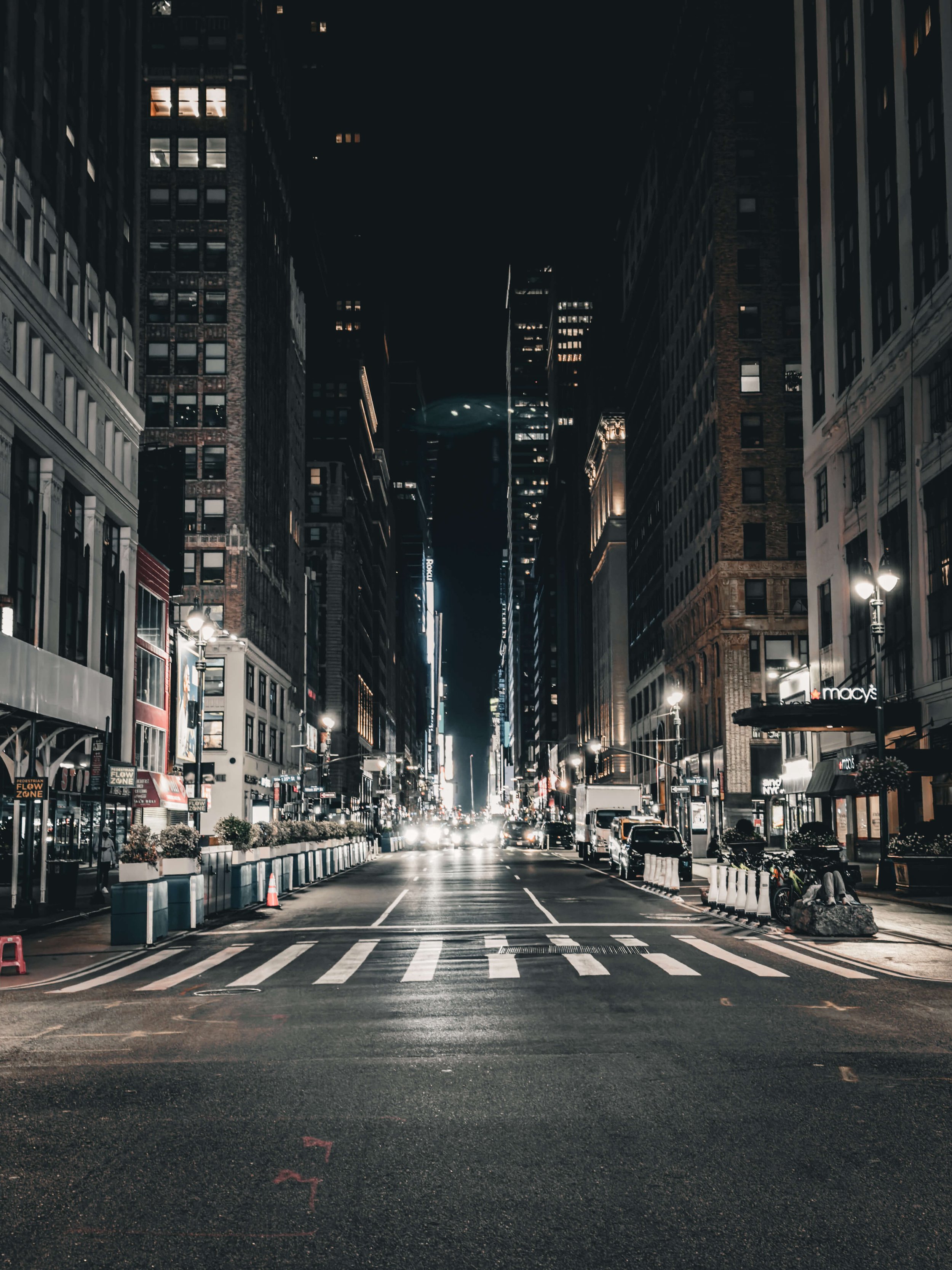 A city street at night with tall buildings, illuminated storefronts, streetlights, and parked cars.