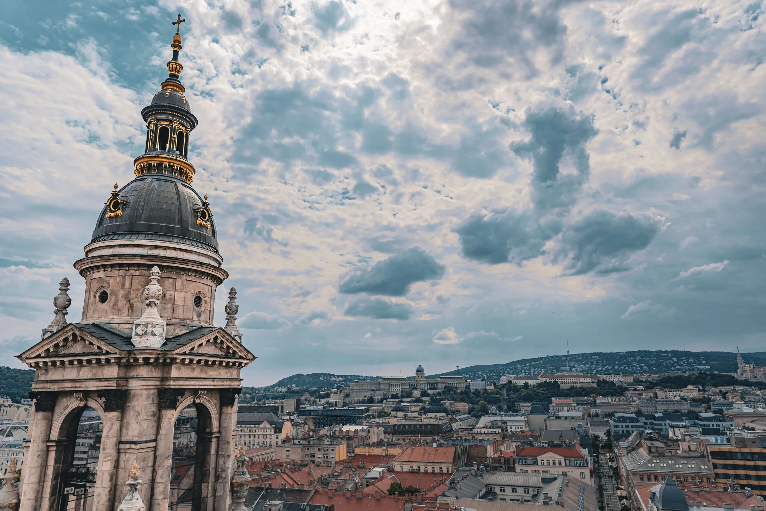 A historic building with a decorative dome and cross on top, overlooking a cityscape with numerous rooftops and a distant hill with buildings and trees under a partly cloudy sky.