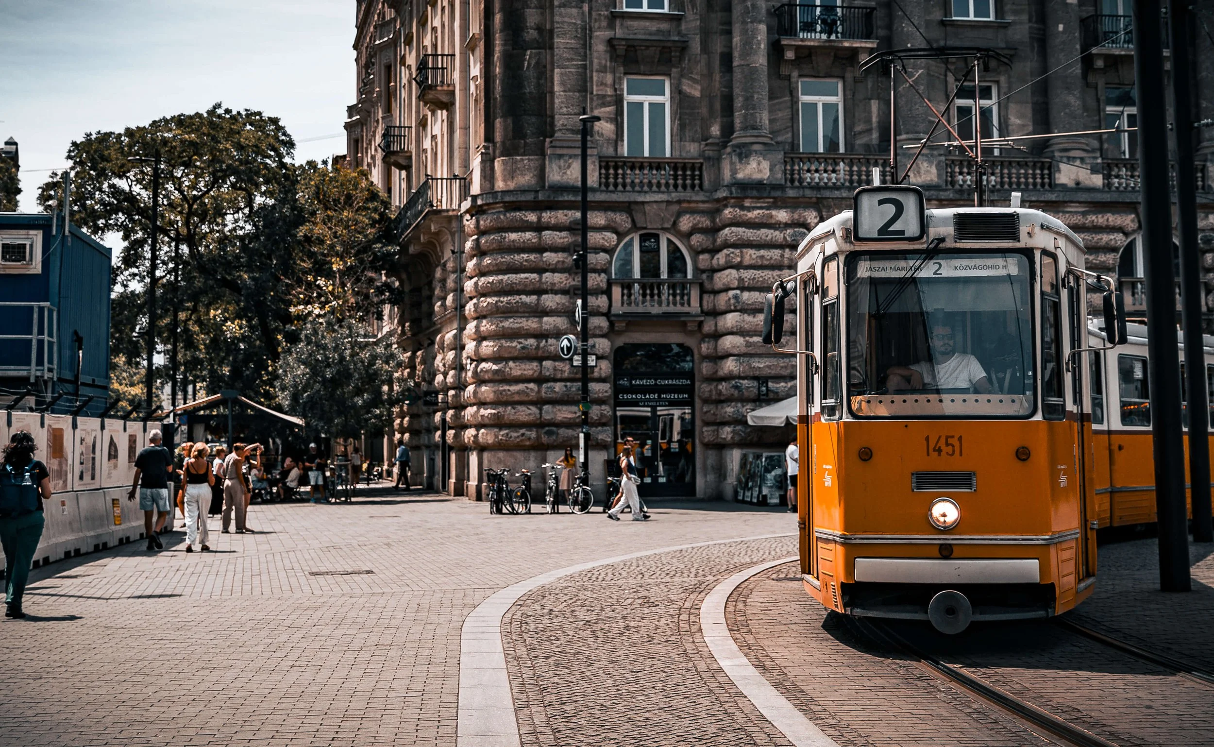 A yellow tram on city street with people walking, bicycles parked, historic building with rounded stone corners, and trees in the background.