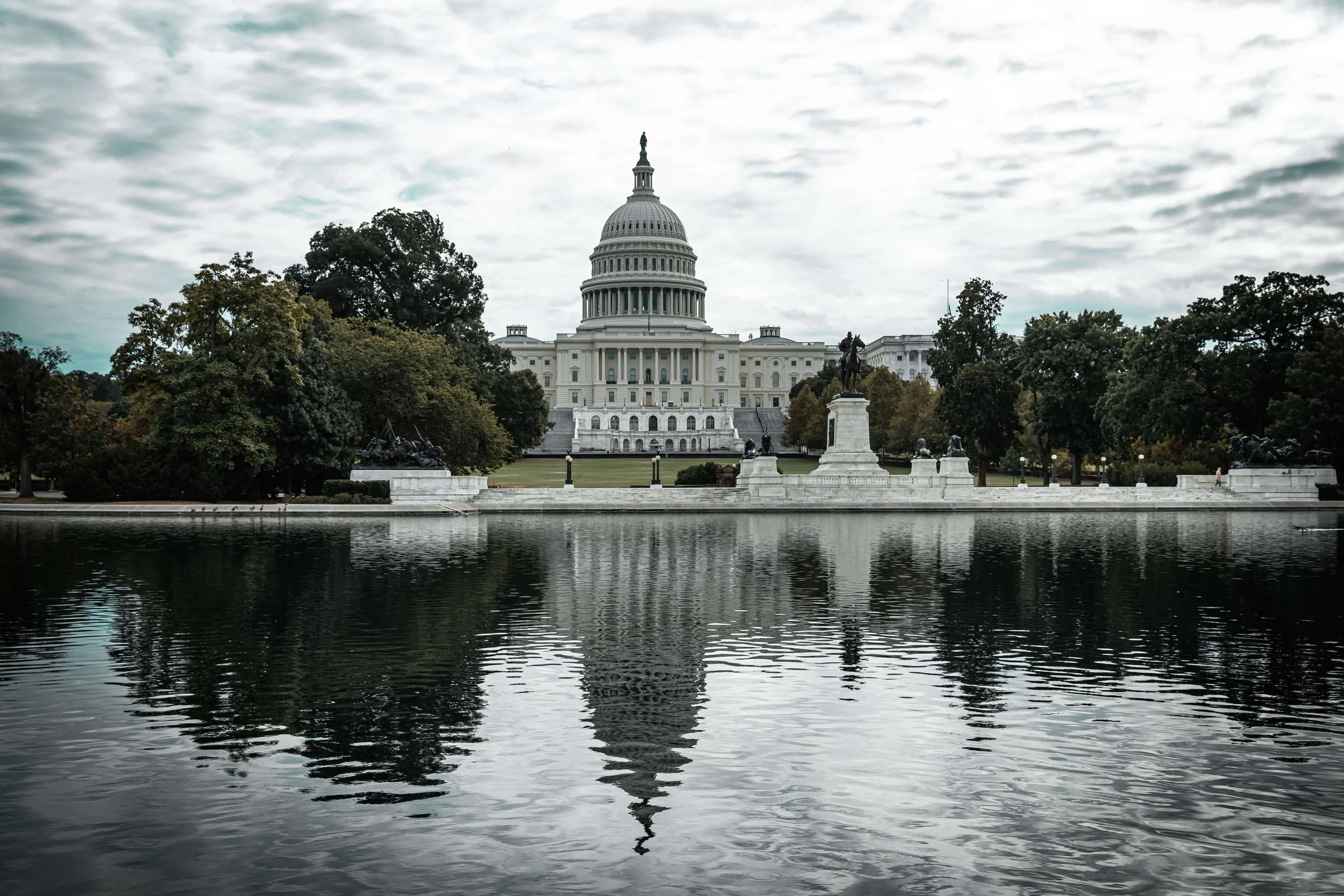 The United States Capitol building in Washington, D.C., viewed from across a reflecting pool with trees on either side, under a cloudy sky.