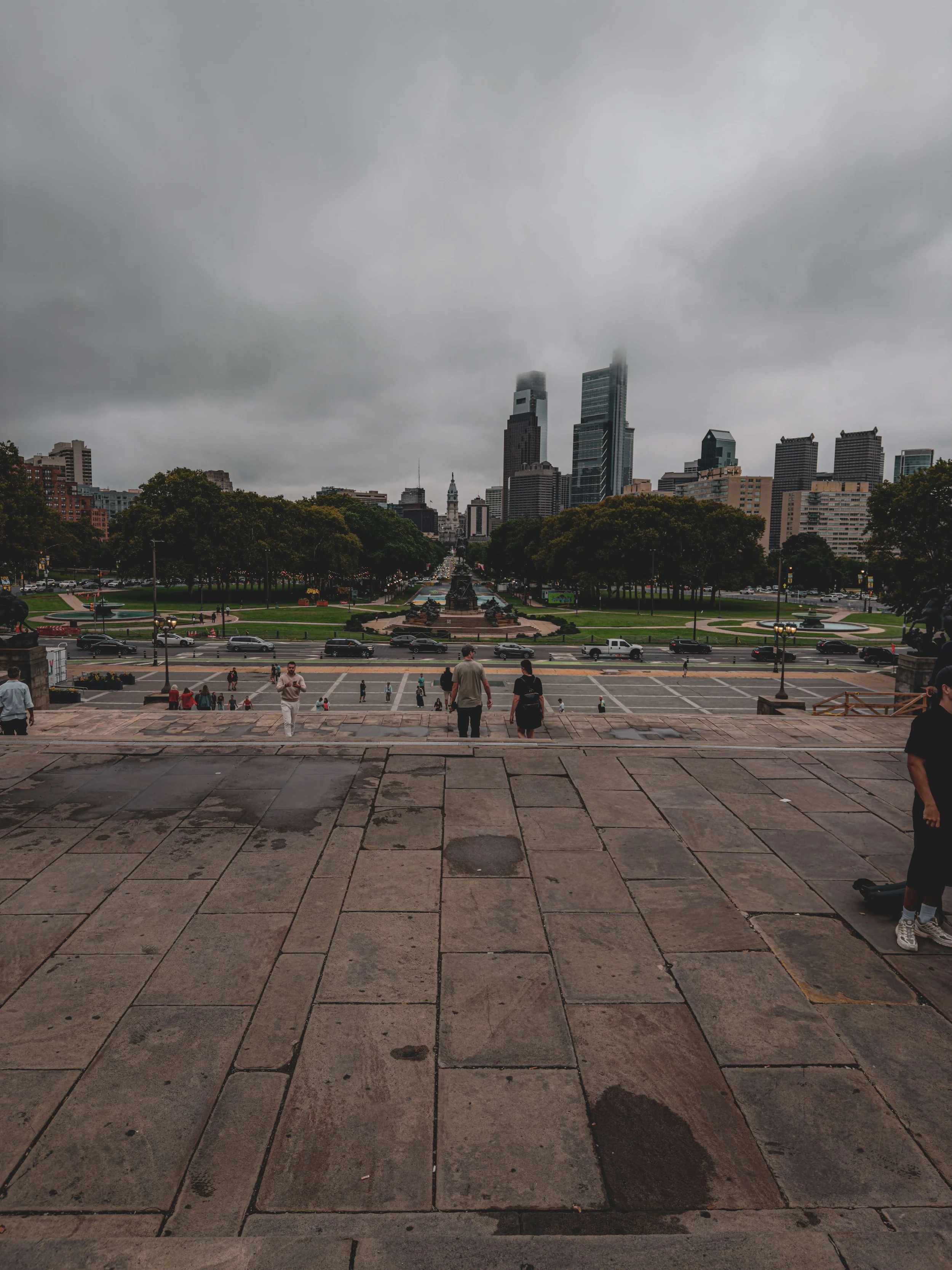 City skyline with tall buildings in the distance, viewed from steps with people walking downward on a cloudy day.