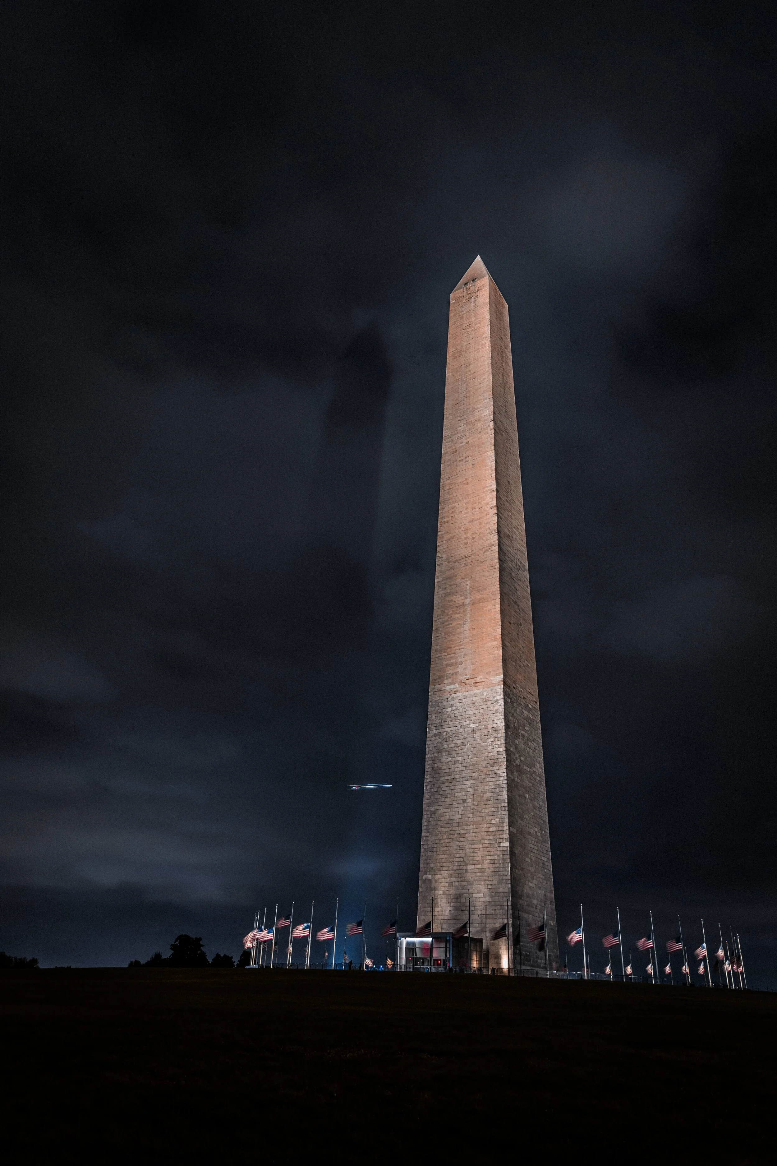 Nighttime view of the Washington Monument in Washington D.C., illuminated, with a dark cloudy sky in the background and American flags surrounding the base.