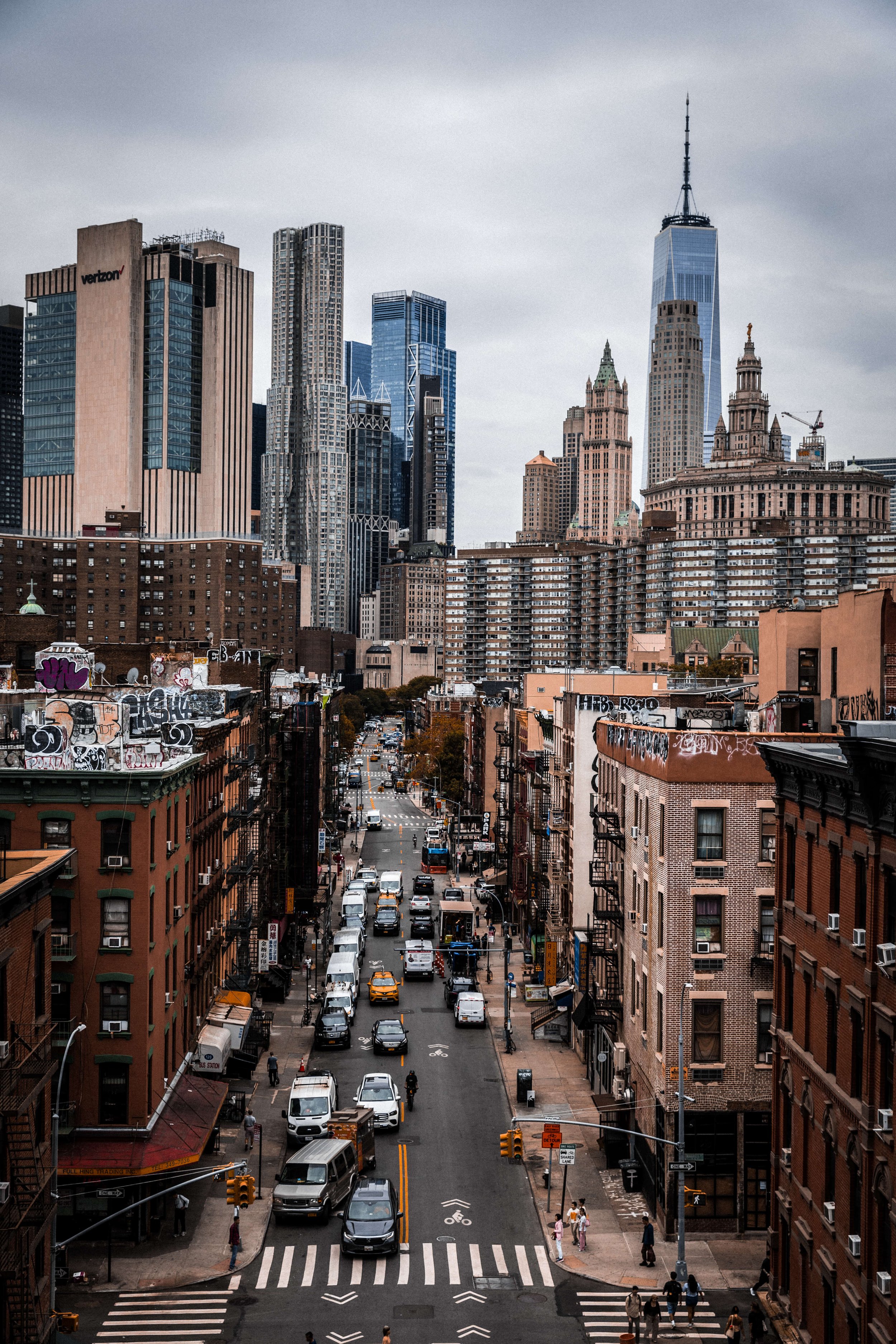 Downtown New York City street scene with tall skyscrapers, including the One World Trade Center, overcast sky, cars and pedestrians on the street.