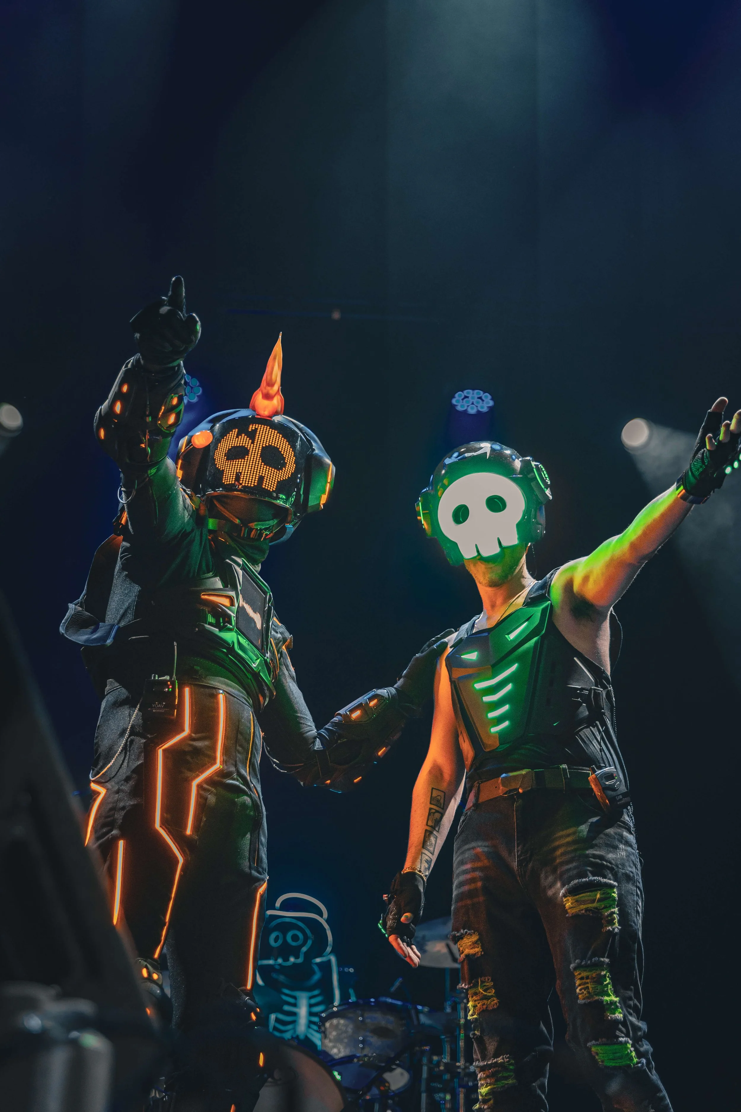 Two individuals wearing illuminated neon costumes and skull masks, celebrating or performing on a dark stage surrounded by stage lights and equipment.