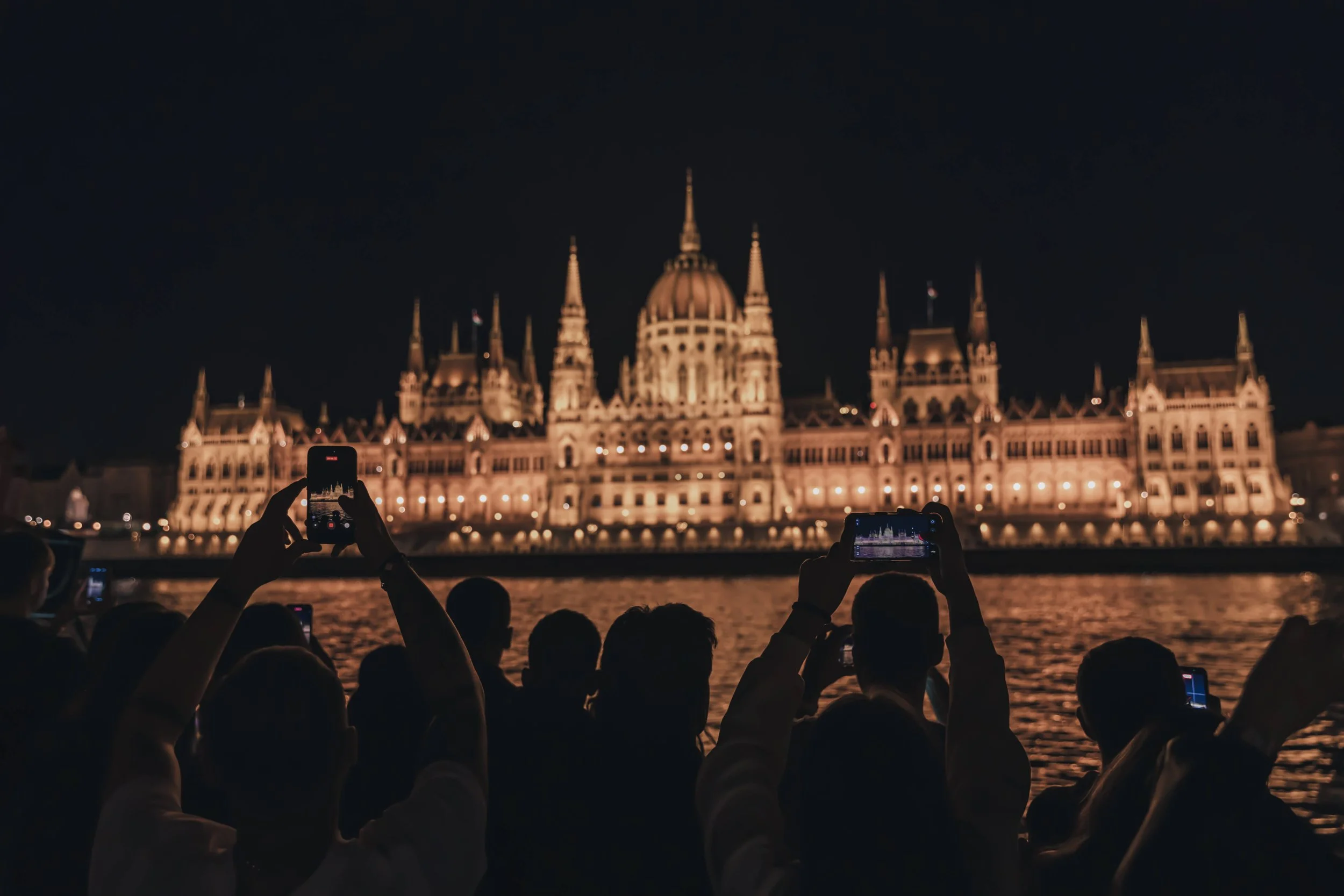 Nighttime view of the Hungarian Parliament Building illuminated, with people in the foreground taking photos of it along the riverbank.