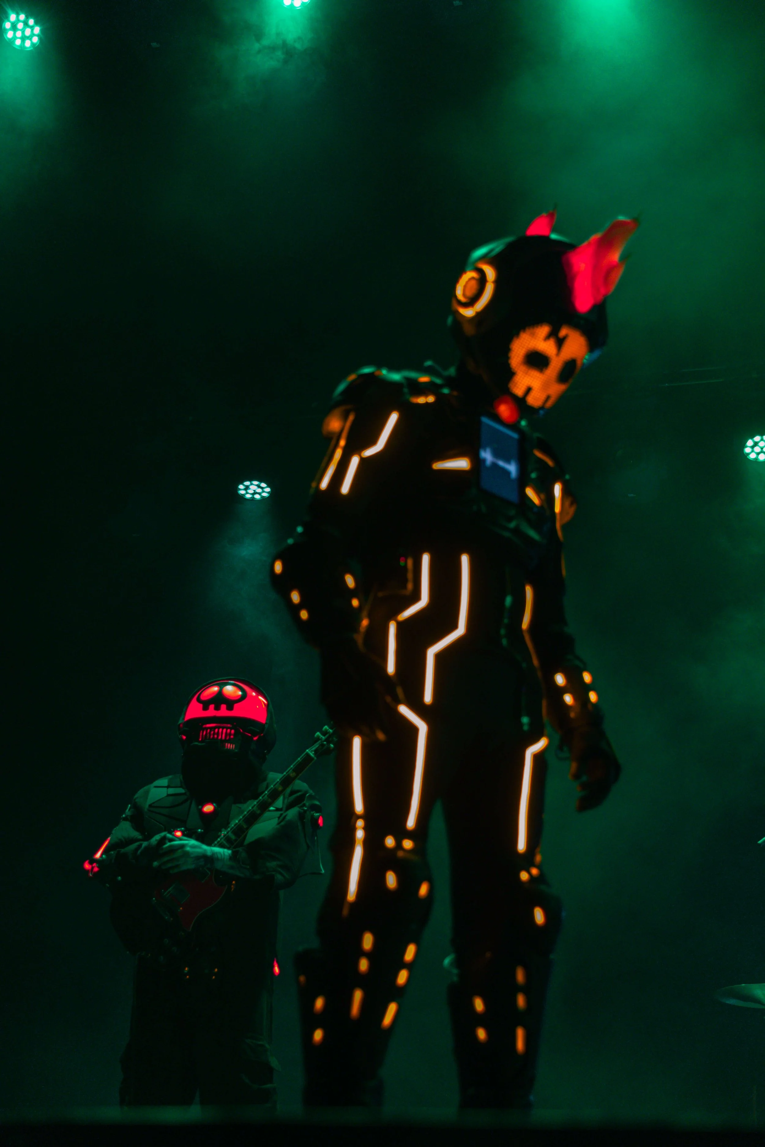 Two performers on stage wearing costumes with LED lights, against a dark background with green stage lighting, one playing a guitar and the other standing, both with skull-themed helmets.