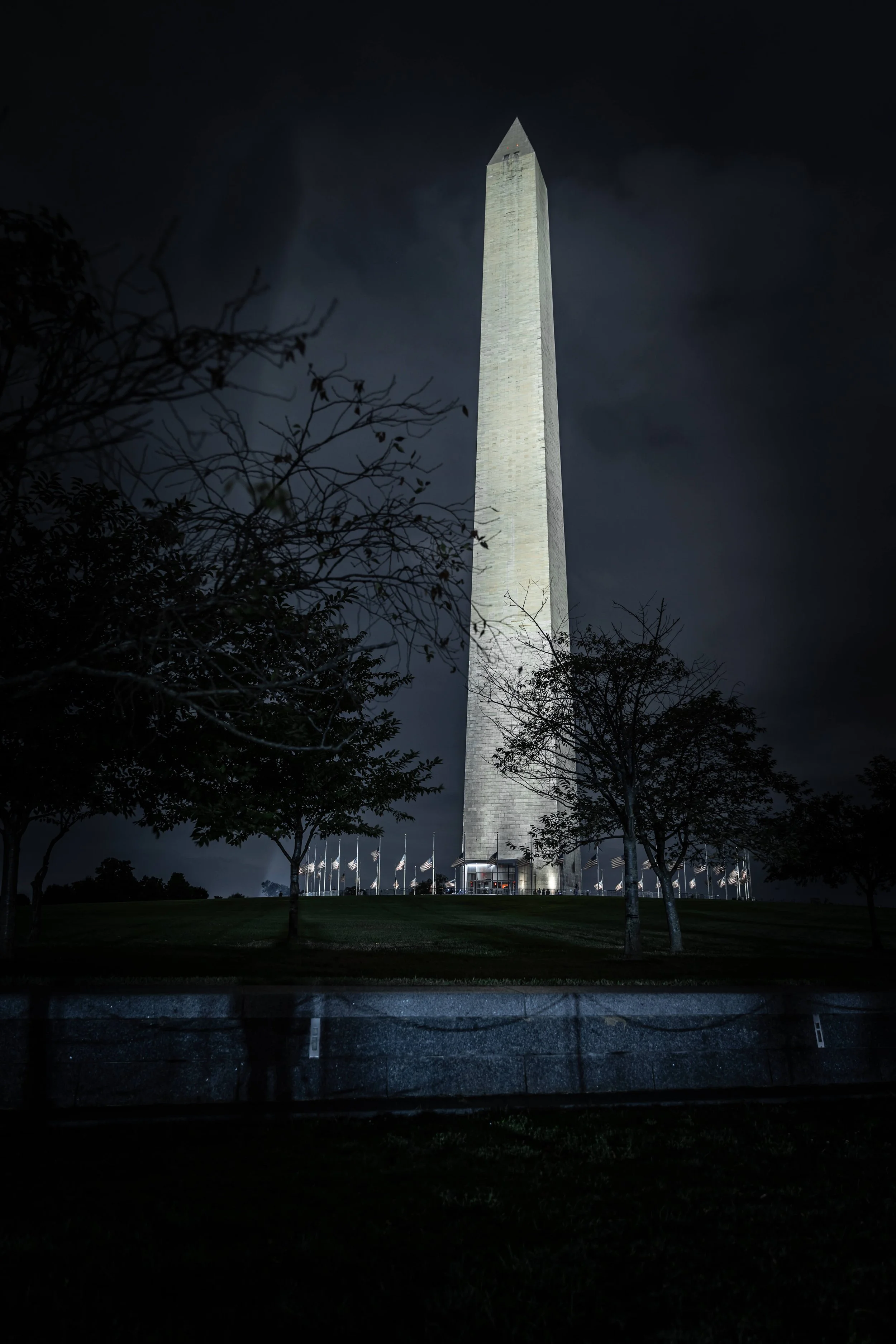 Nighttime photo of the Washington Monument, an obelisk, illuminated against a dark sky, with trees and flagpoles surrounding the base.