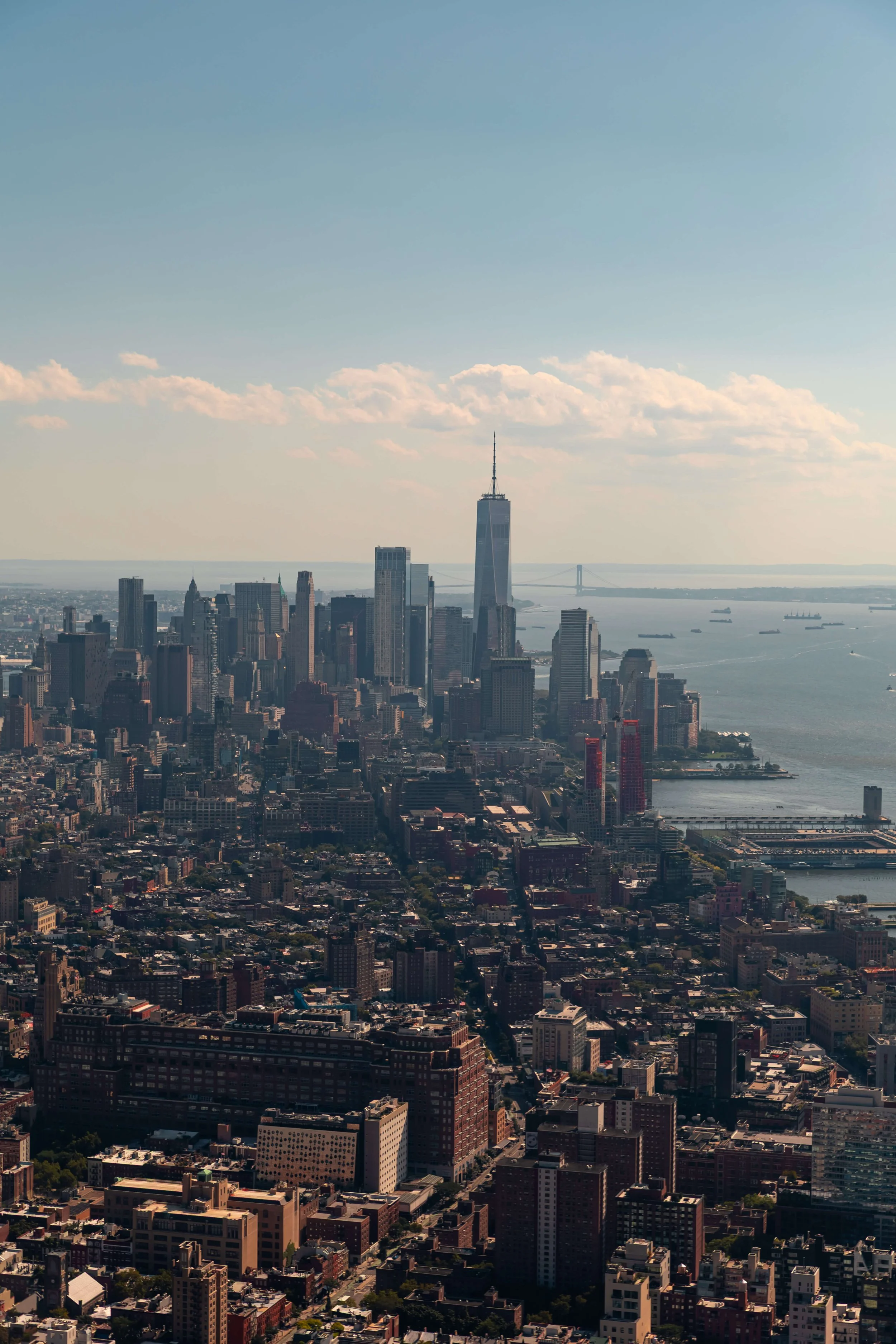 Aerial view of Manhattan skyline featuring One World Trade Center, with bridges and water in the background on a clear day.