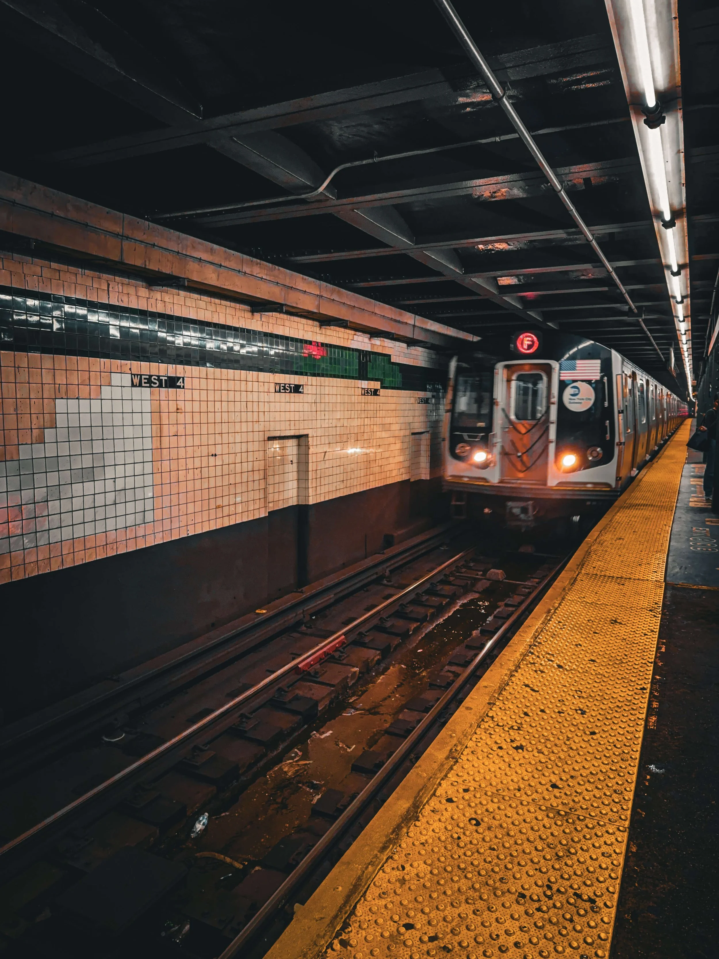 NYC subway train arriving at an underground station platform with yellow tactile paving, black ceiling, tiled wall, and passengers waiting.