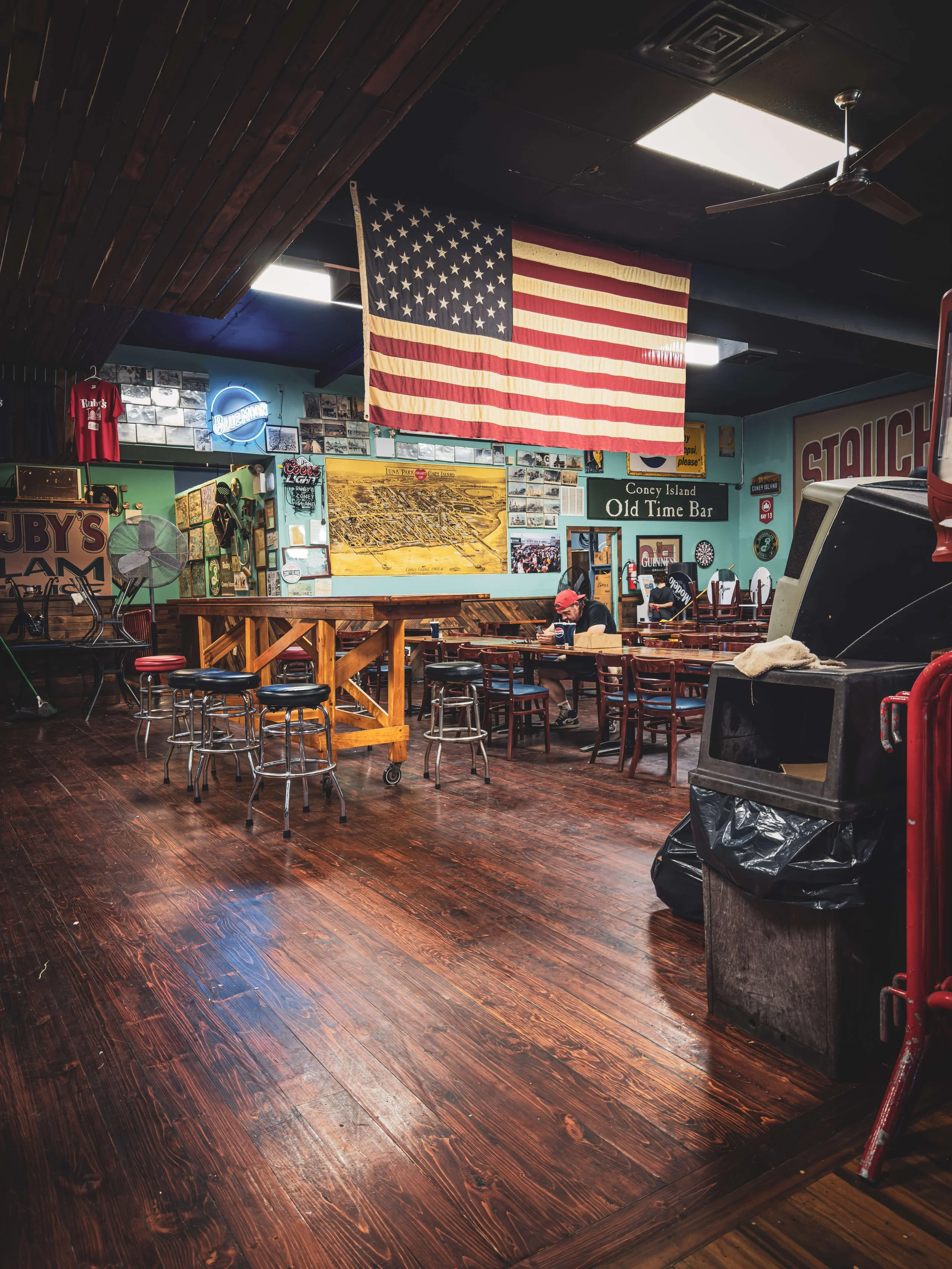 Interior of a bar or restaurant with a large American flag hanging from the ceiling, wooden floors, and various wall decorations including signs and photographs. There are tables with chairs, some with customers, and a dartboard.