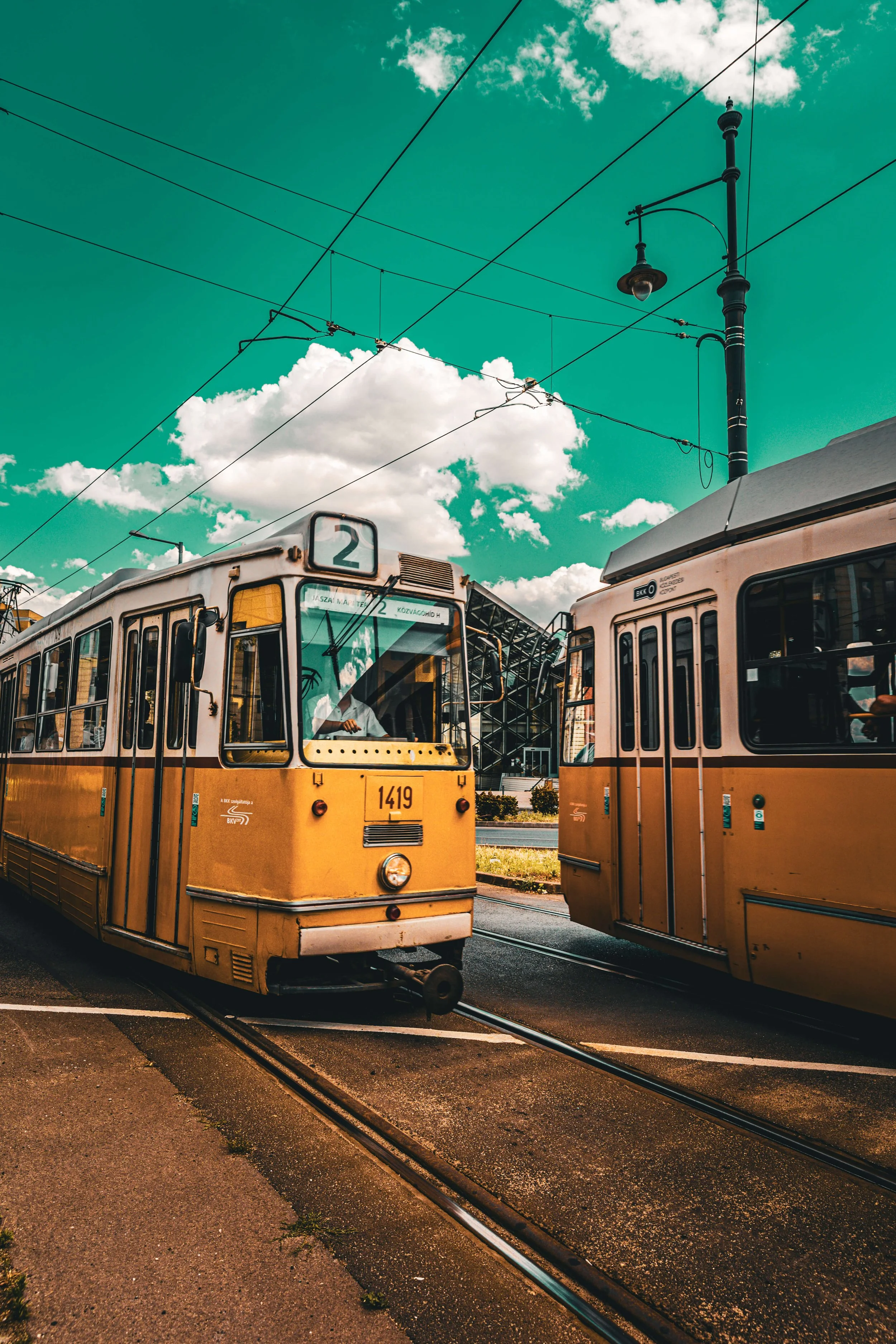 Two yellow trams on tracks under a blue sky with clouds, overhead power lines, and a lamppost.