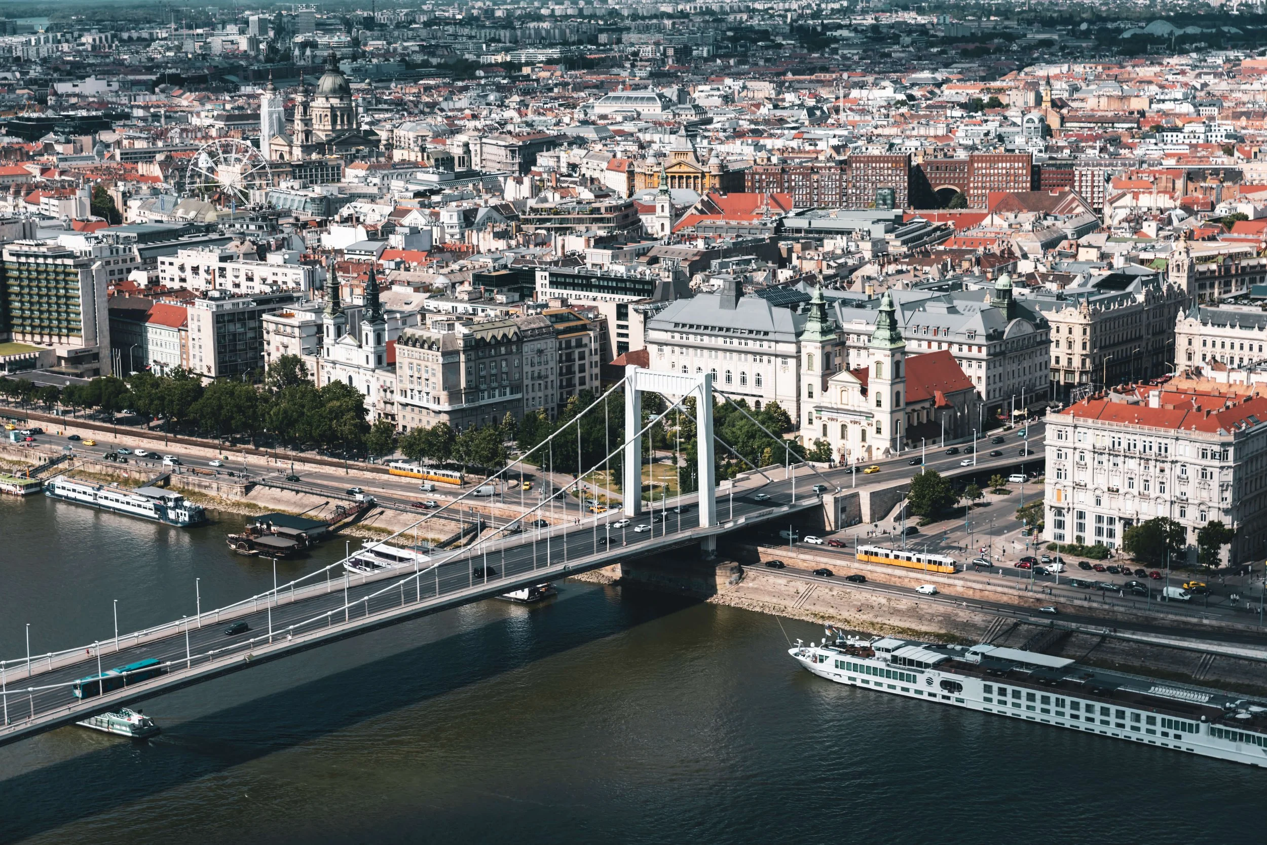Aerial view of a city with a river, numerous buildings, a bridge, a Ferris wheel, and boats on the water.