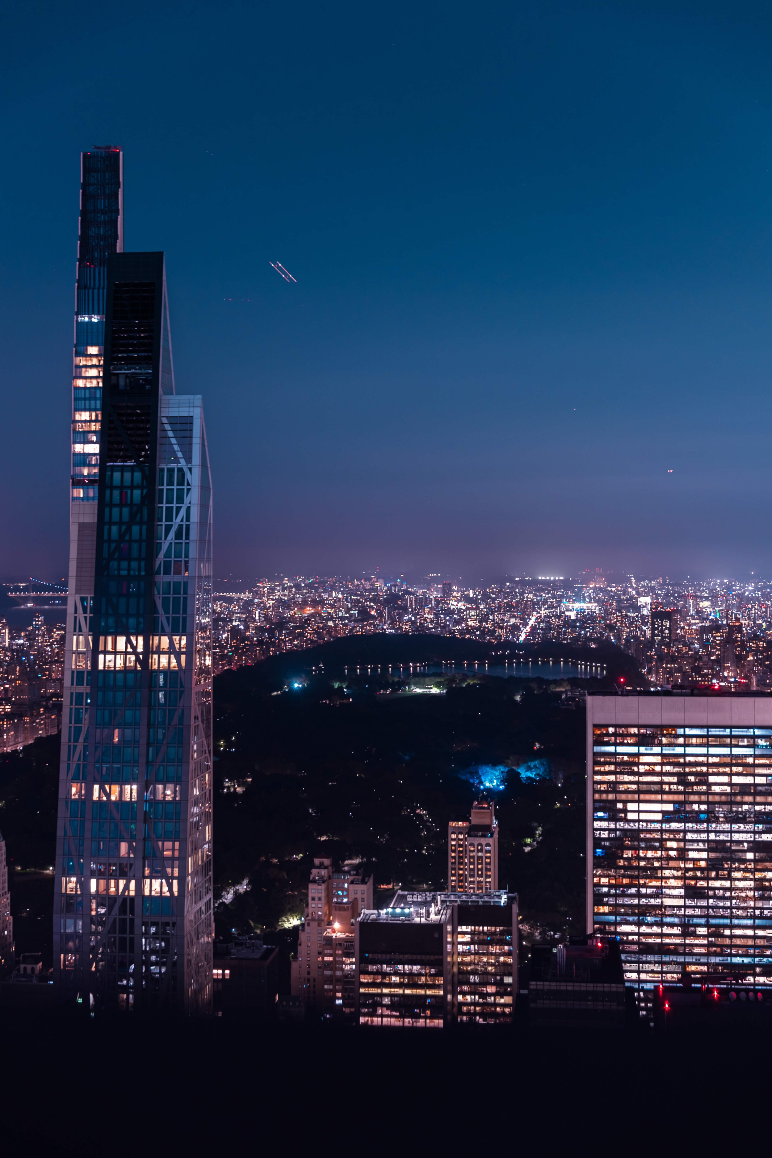 Nighttime cityscape with tall modern skyscrapers, including a distinctive twisted glass building, illuminated windows, and a dark hill with a pond in the distance.