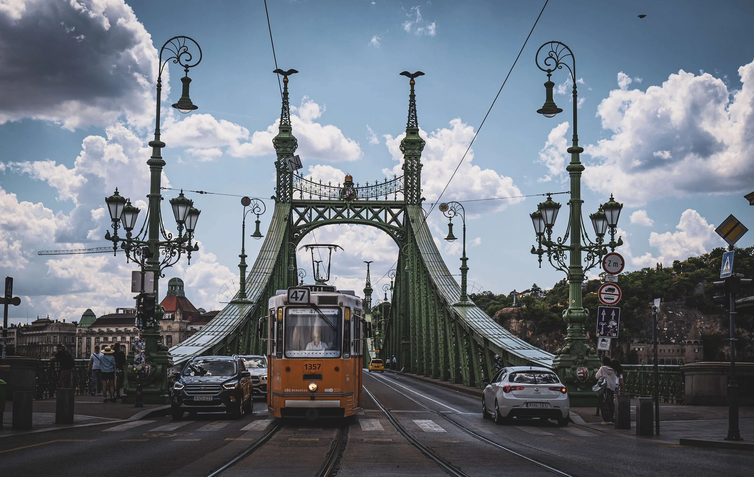 View of a bridge with decorative street lamps and trams in a city, with people walking and cars on the street and a partly cloudy sky in the background.