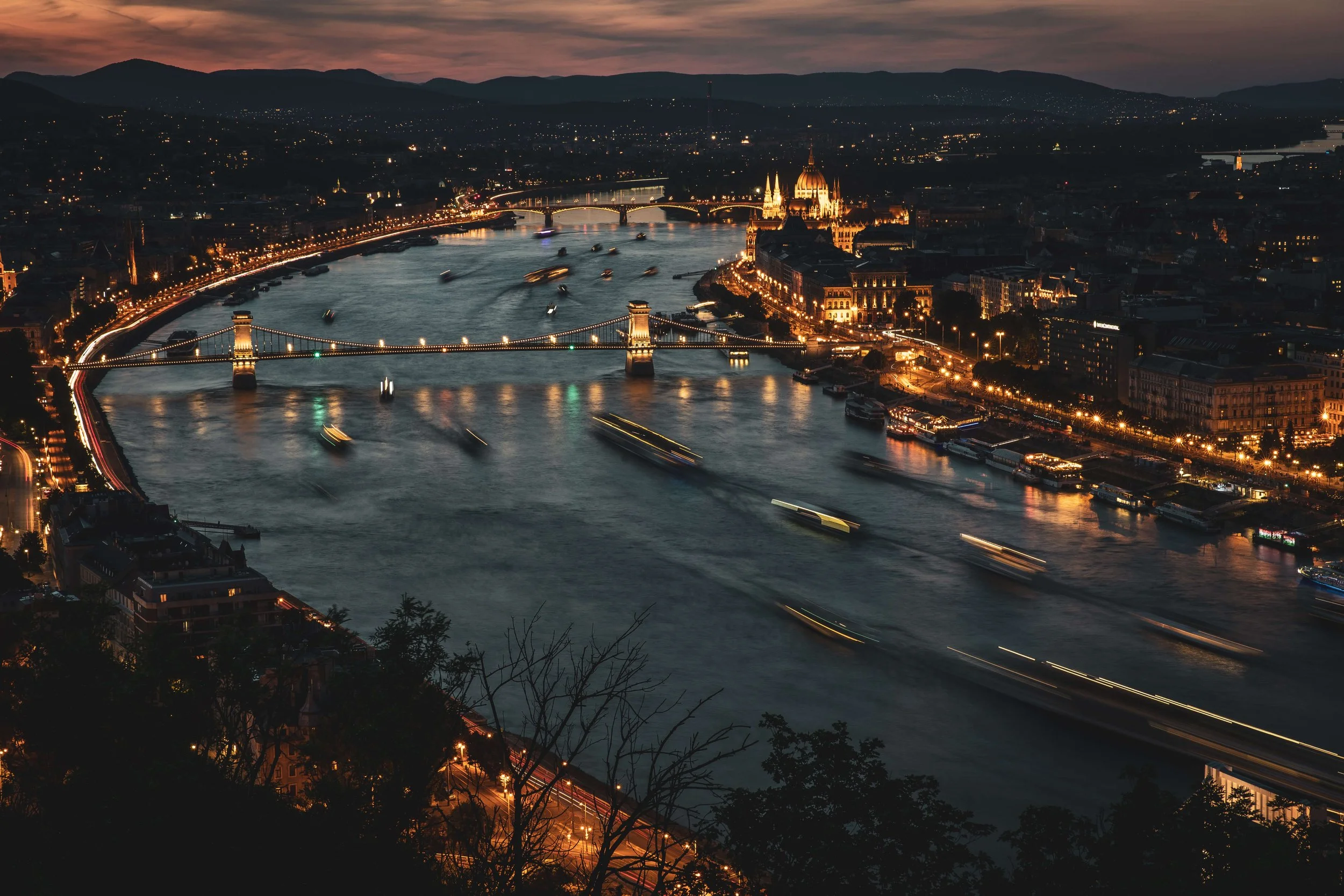 Nighttime view of a city with illuminated bridges crossing a river, historic buildings, and mountains in the background.