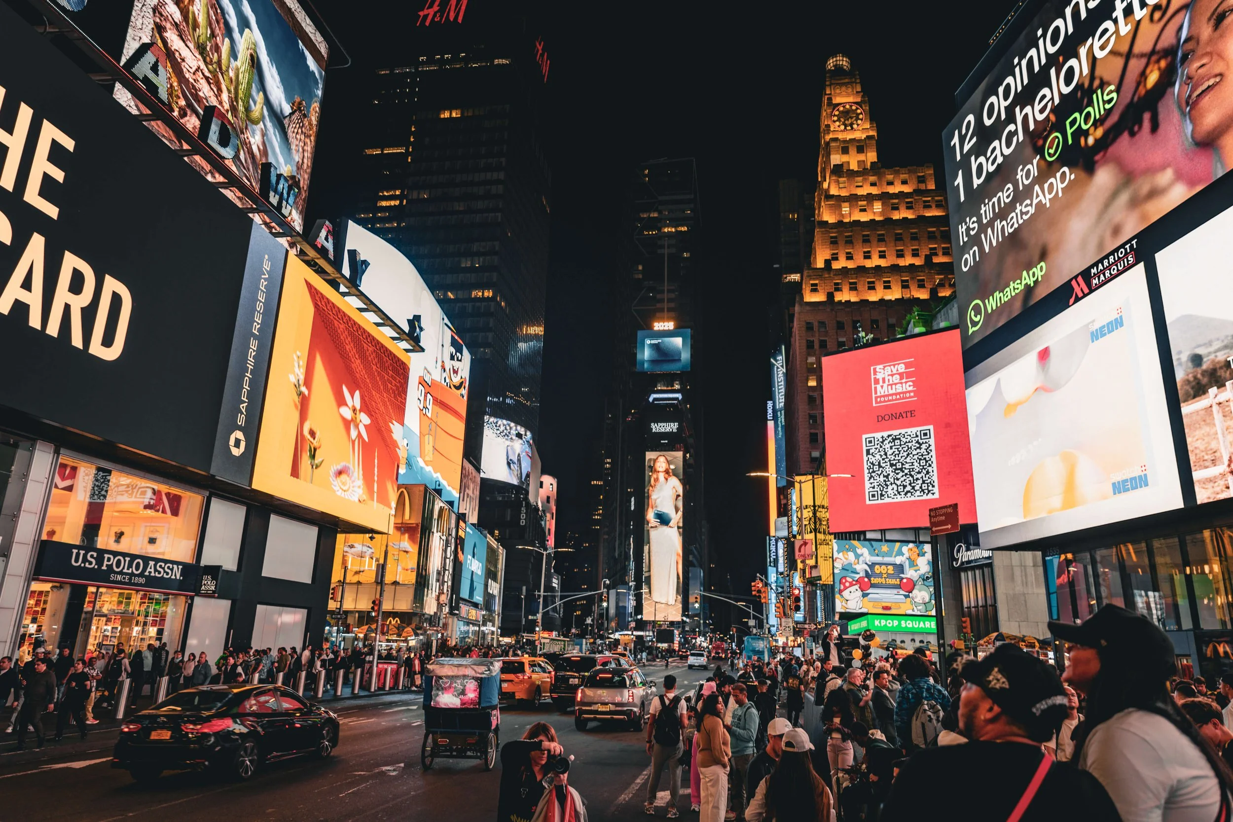 Busy city street at night with bright digital billboards and advertisements, including a Samsung logo, a Save the Music Foundation QR code, and many pedestrians and cars.