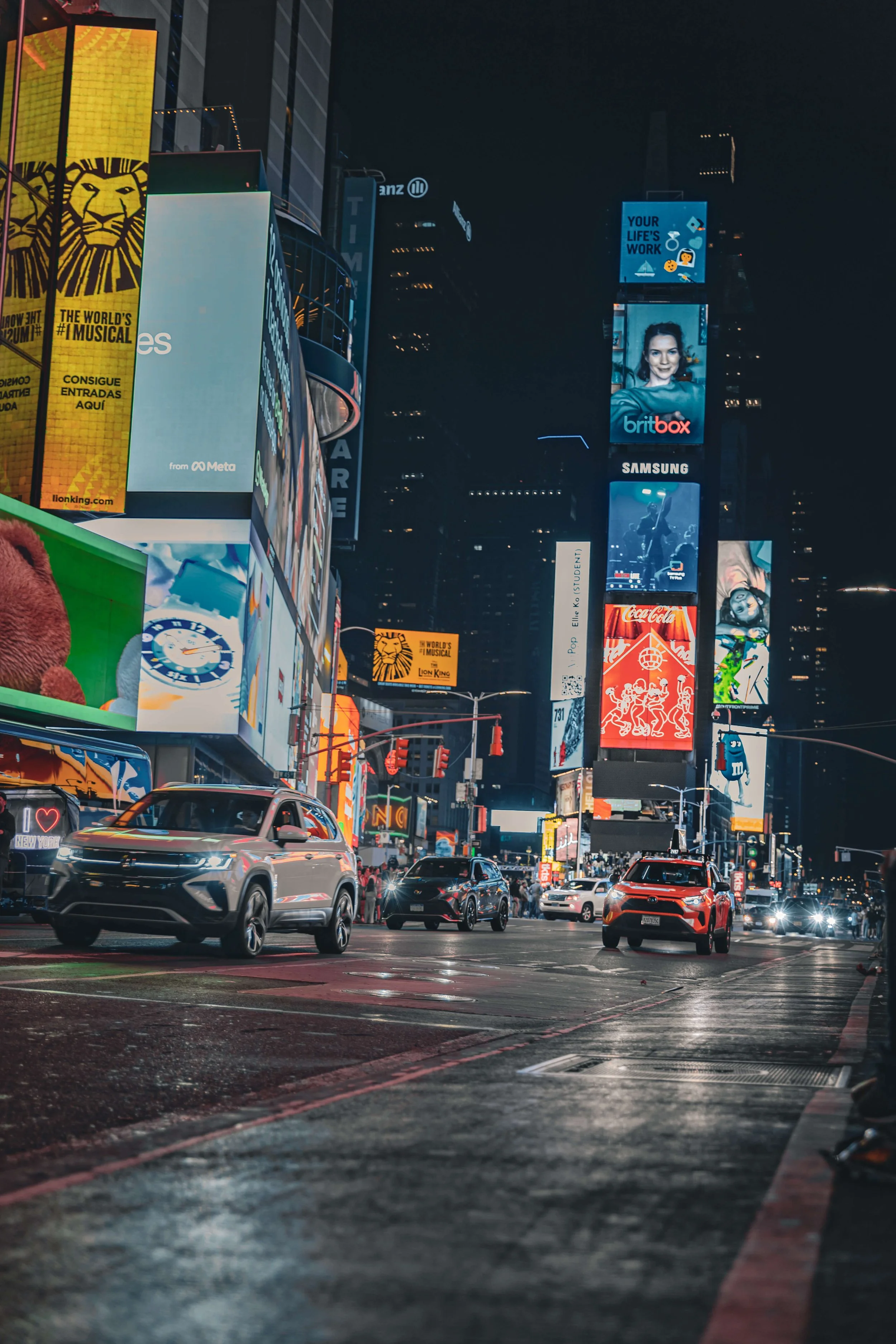 A city street scene at night with illuminated billboards and advertisements, cars driving through the intersection, and skyscrapers in the background.