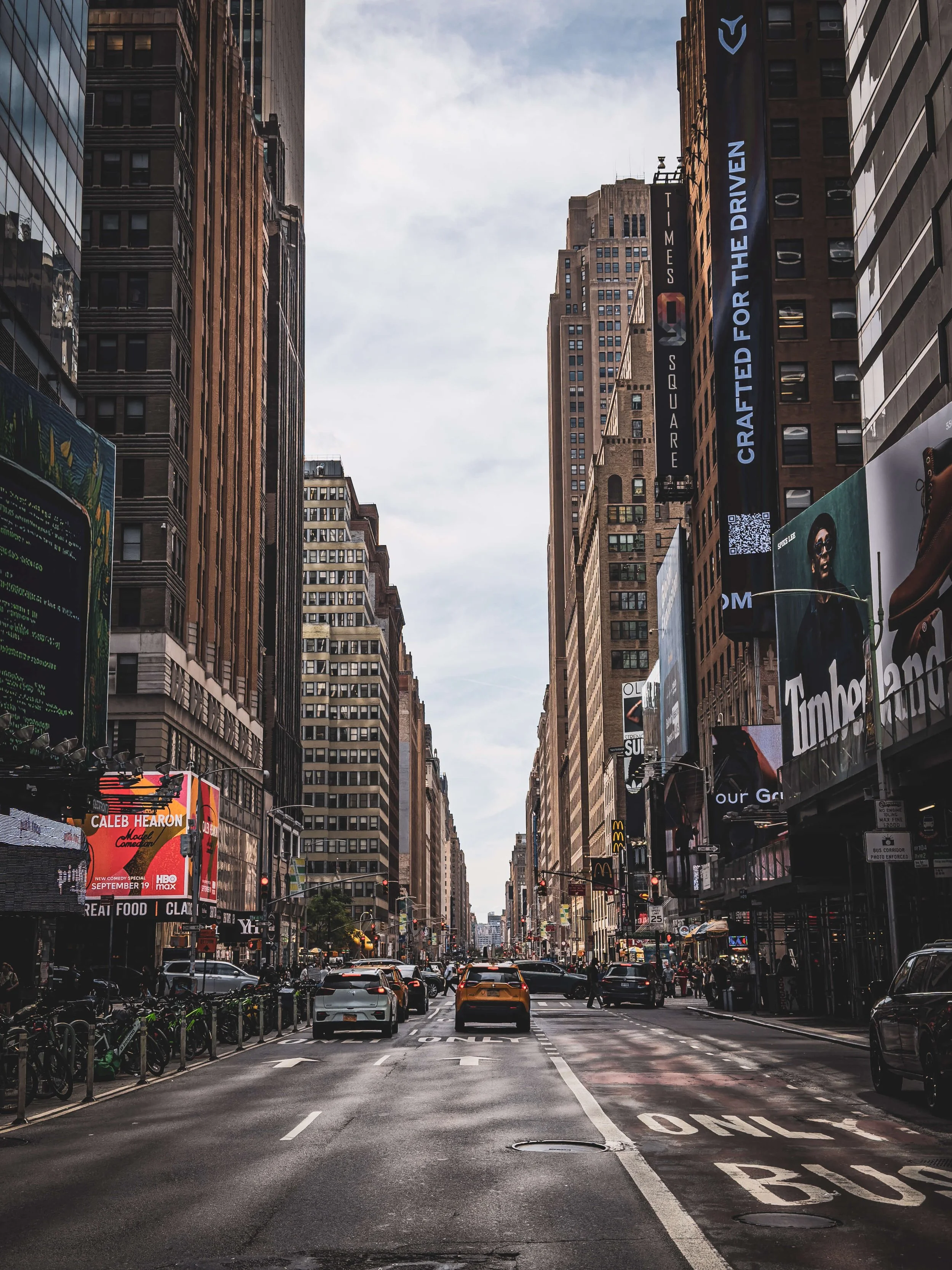 Urban city street with tall buildings, cars, bicycles, and billboards, with cloudy sky above.