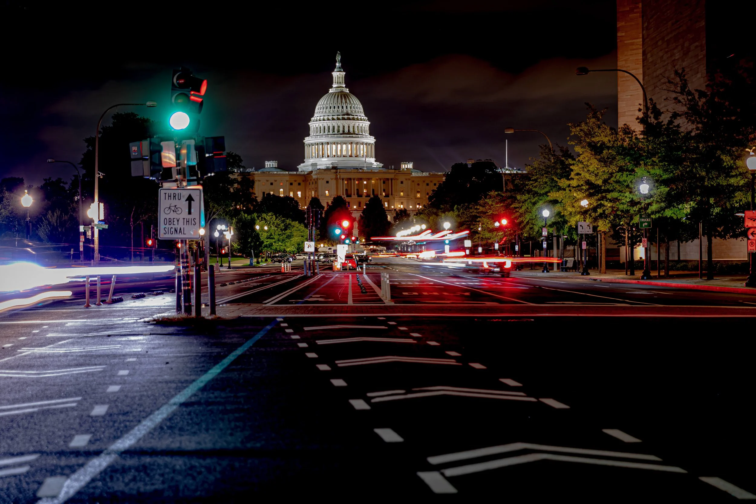 Nighttime view of the U.S. Capitol building in Washington, D.C., with light trails of passing cars on the street, traffic lights, and street signs visible, including one that says 'Thru Bicycle Obey This Signal.'