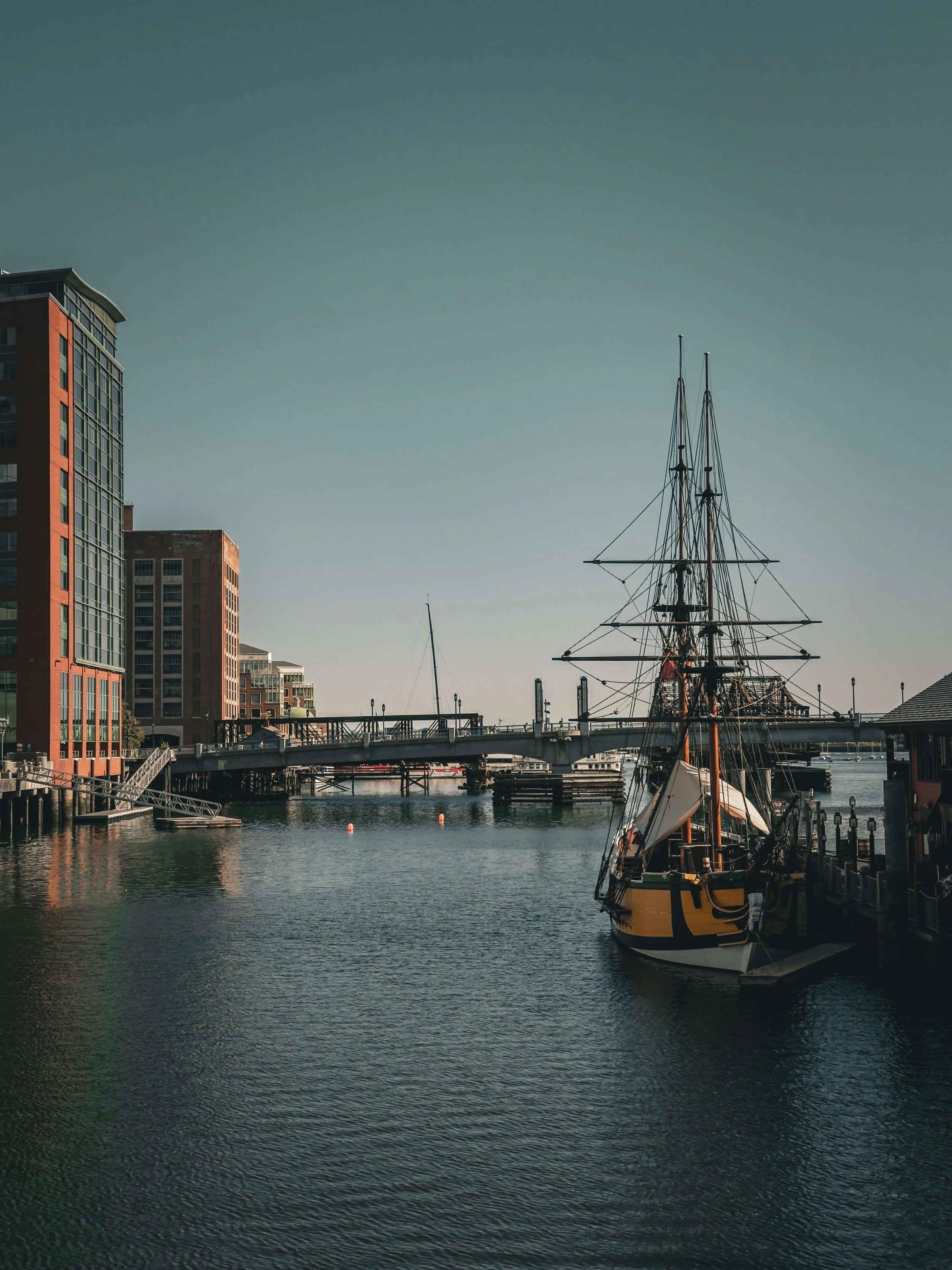 A harbor scene with a historic tall ship docked near modern buildings, a bridge crossing over the water, and clear blue sky.