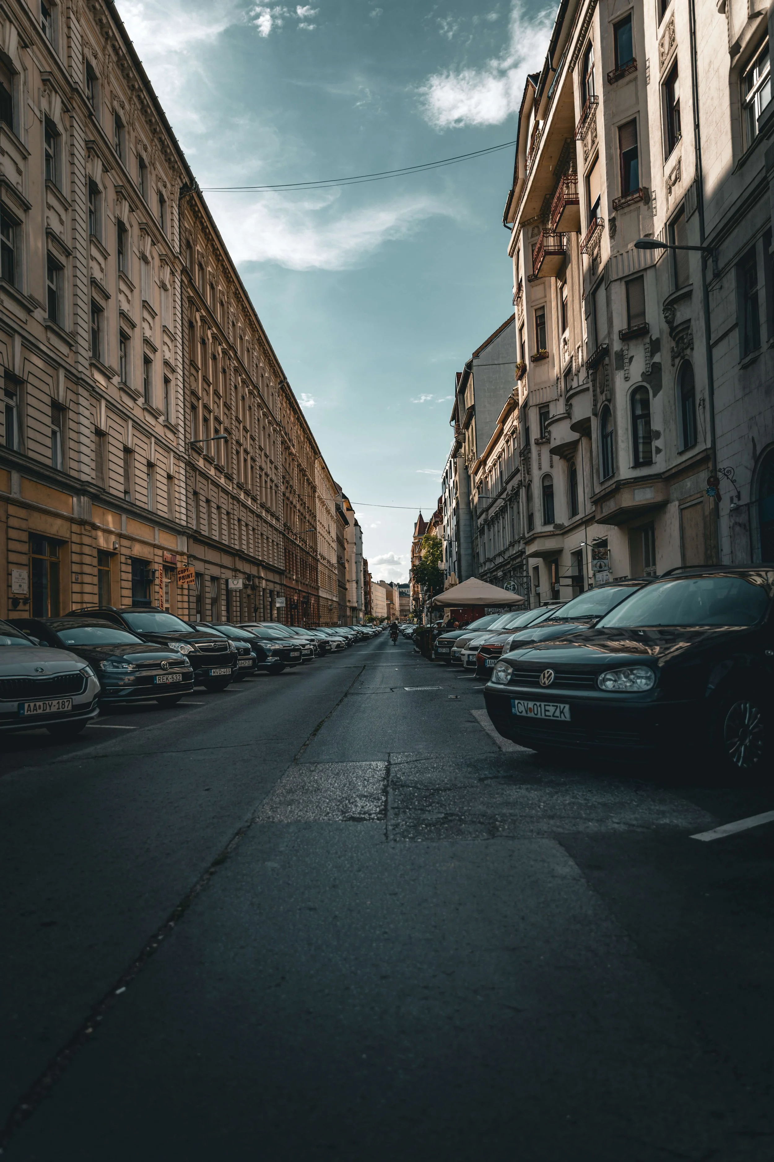 City street with parked cars lining both sides, old buildings with detailed facades, street lamps, and blue sky with clouds overhead.