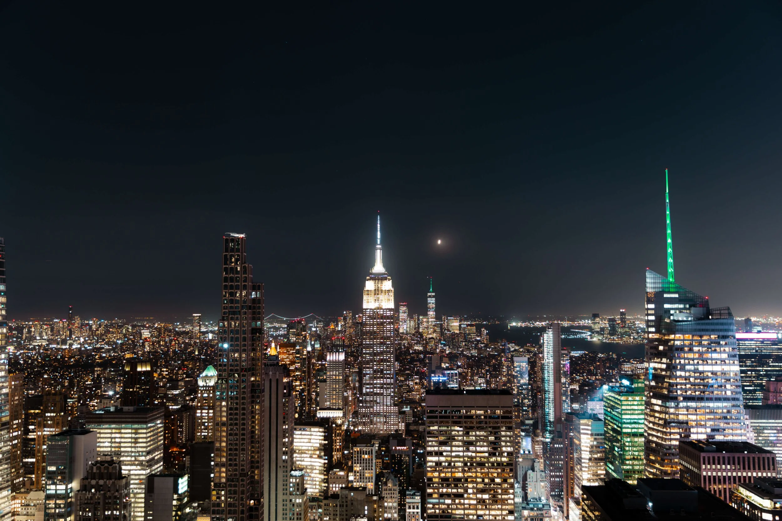 Nighttime city skyline of New York City with illuminated skyscrapers, including the Empire State Building and One World Trade Center, under a dark sky.