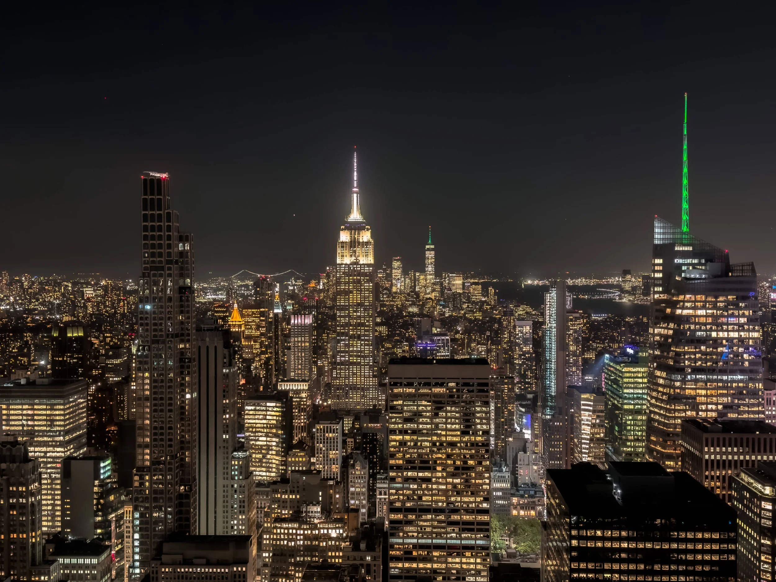 Nighttime cityscape of New York City with illuminated skyscrapers, including the Empire State Building with its spire lit up, and a taller building with a green-lit spire on the right.