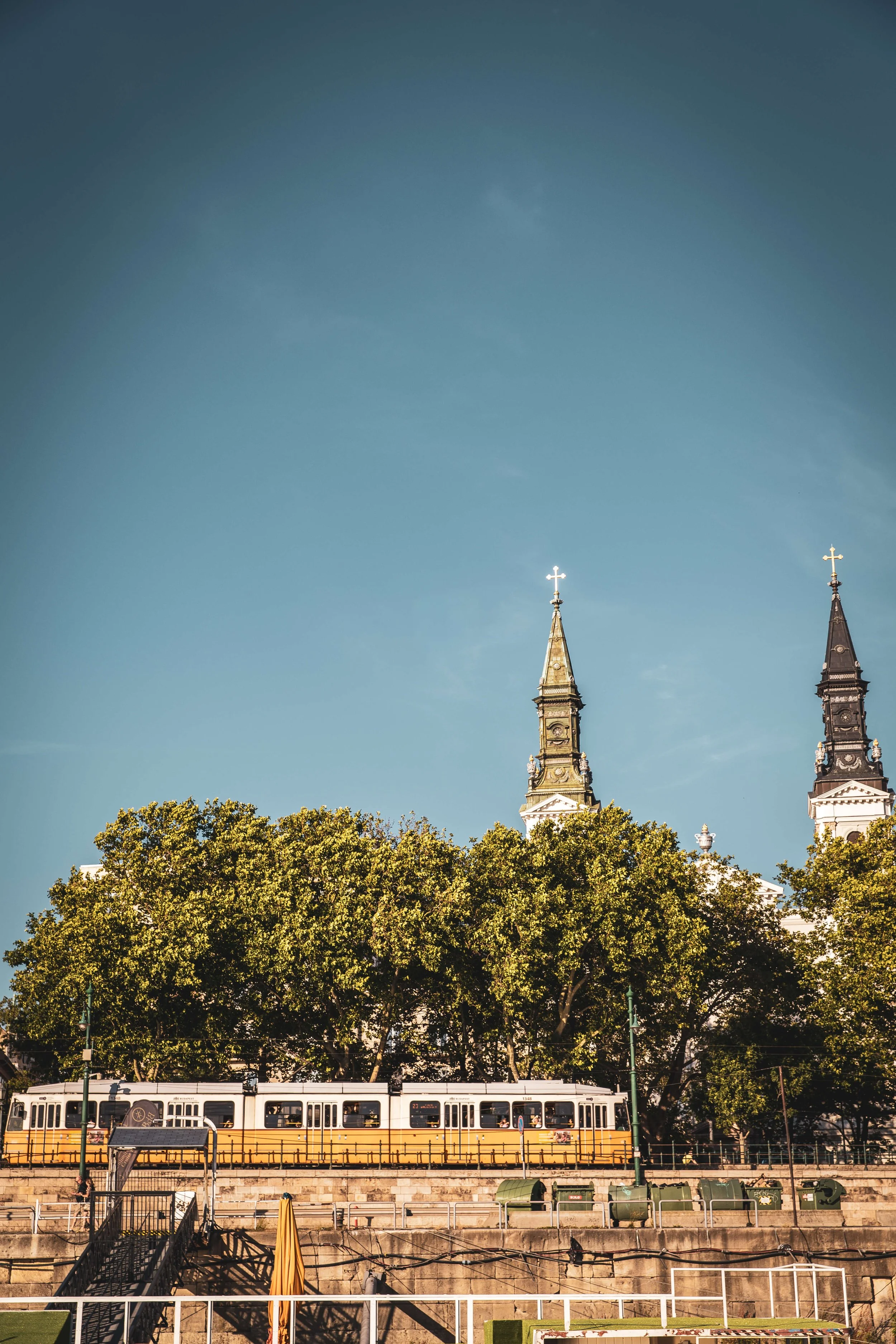A cityscape with historical church towers, green trees in the foreground, a yellow and white tram, and a clear blue sky.