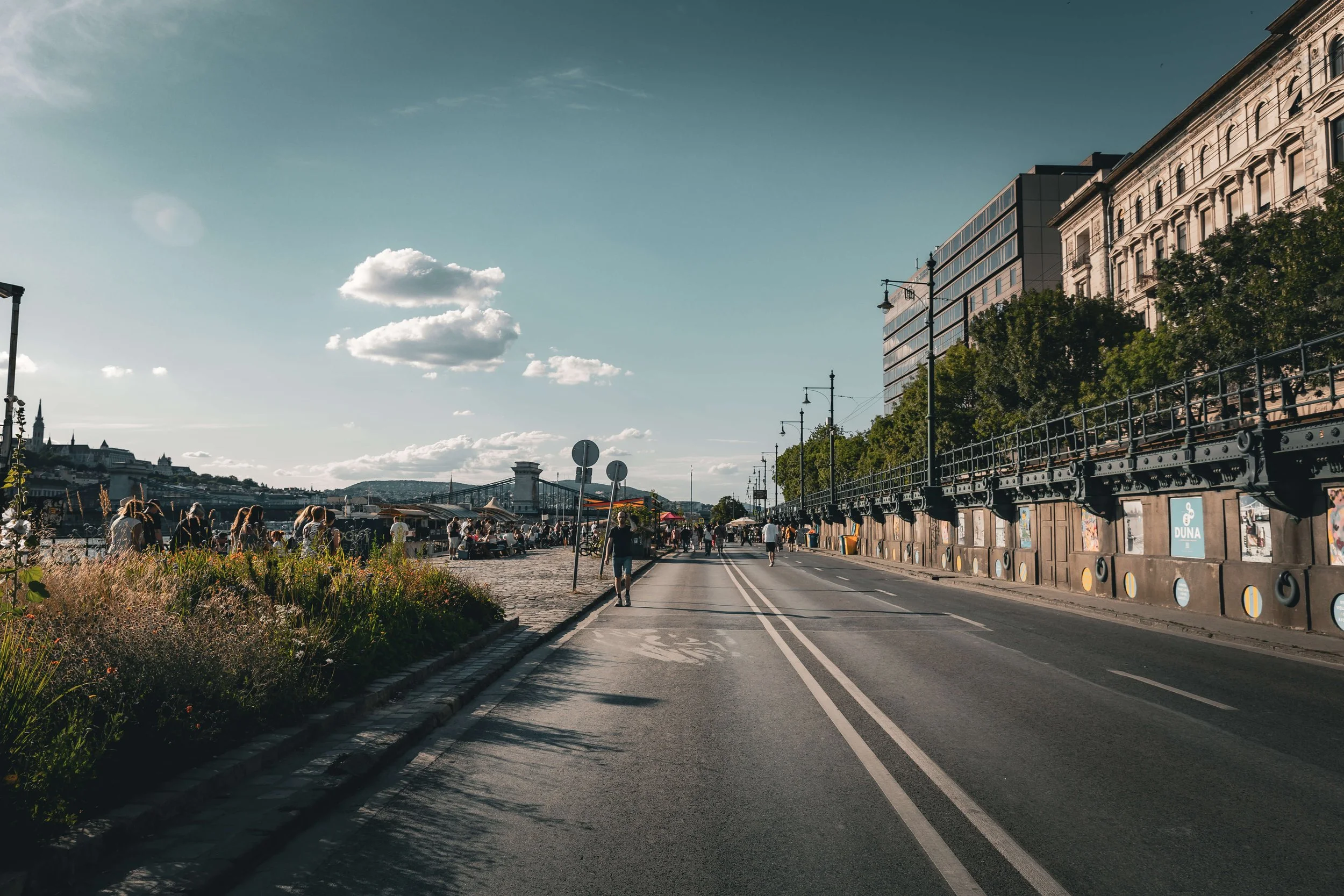 A lively city street scene with people walking along the sidewalk, trees lining the street, and a row of historic and modern buildings. The sky is mostly clear with a few clouds, and the atmosphere suggests late afternoon or early evening.