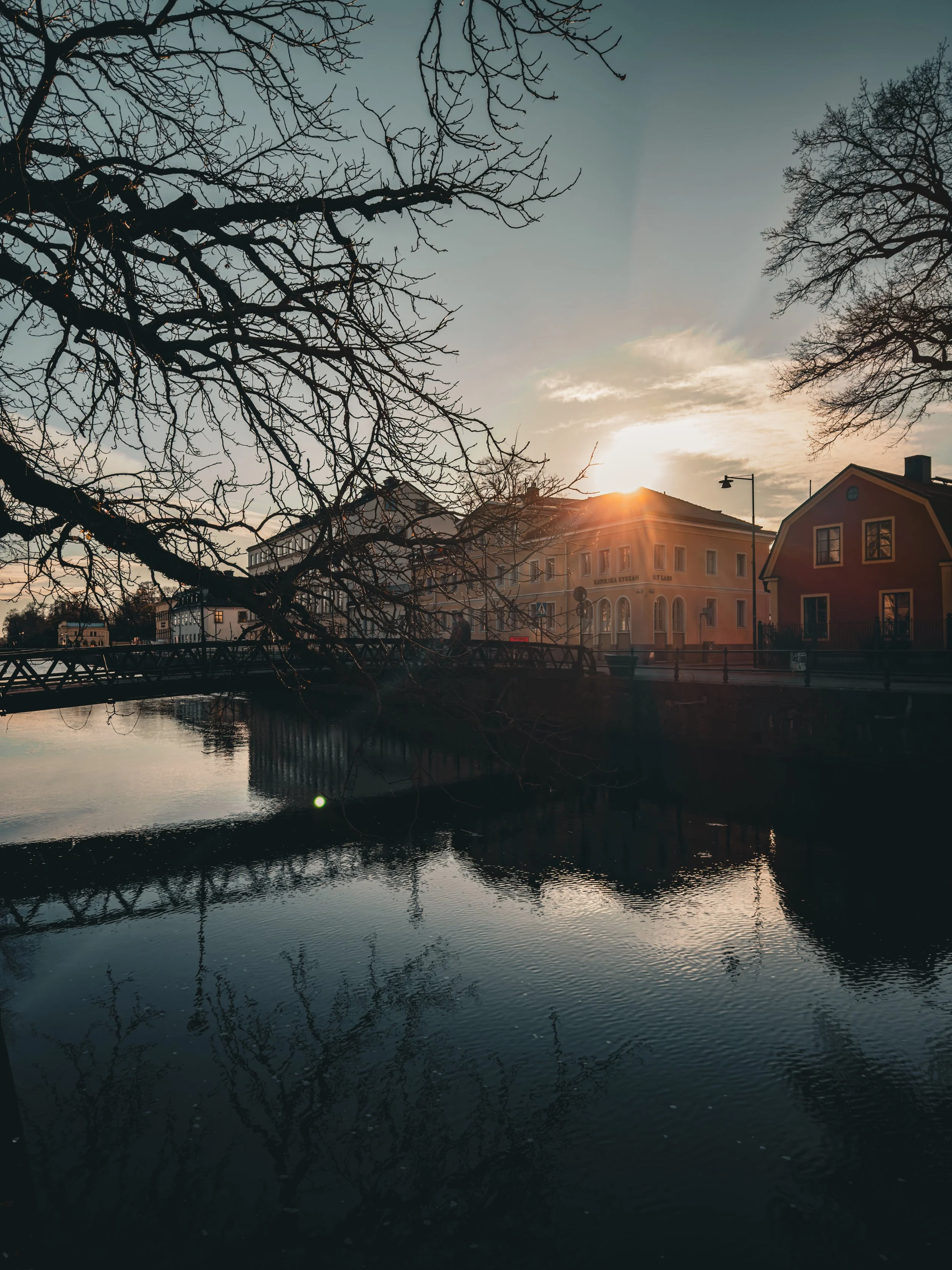 Sunset over historic buildings along a river, with leafless trees and their reflections in the water.