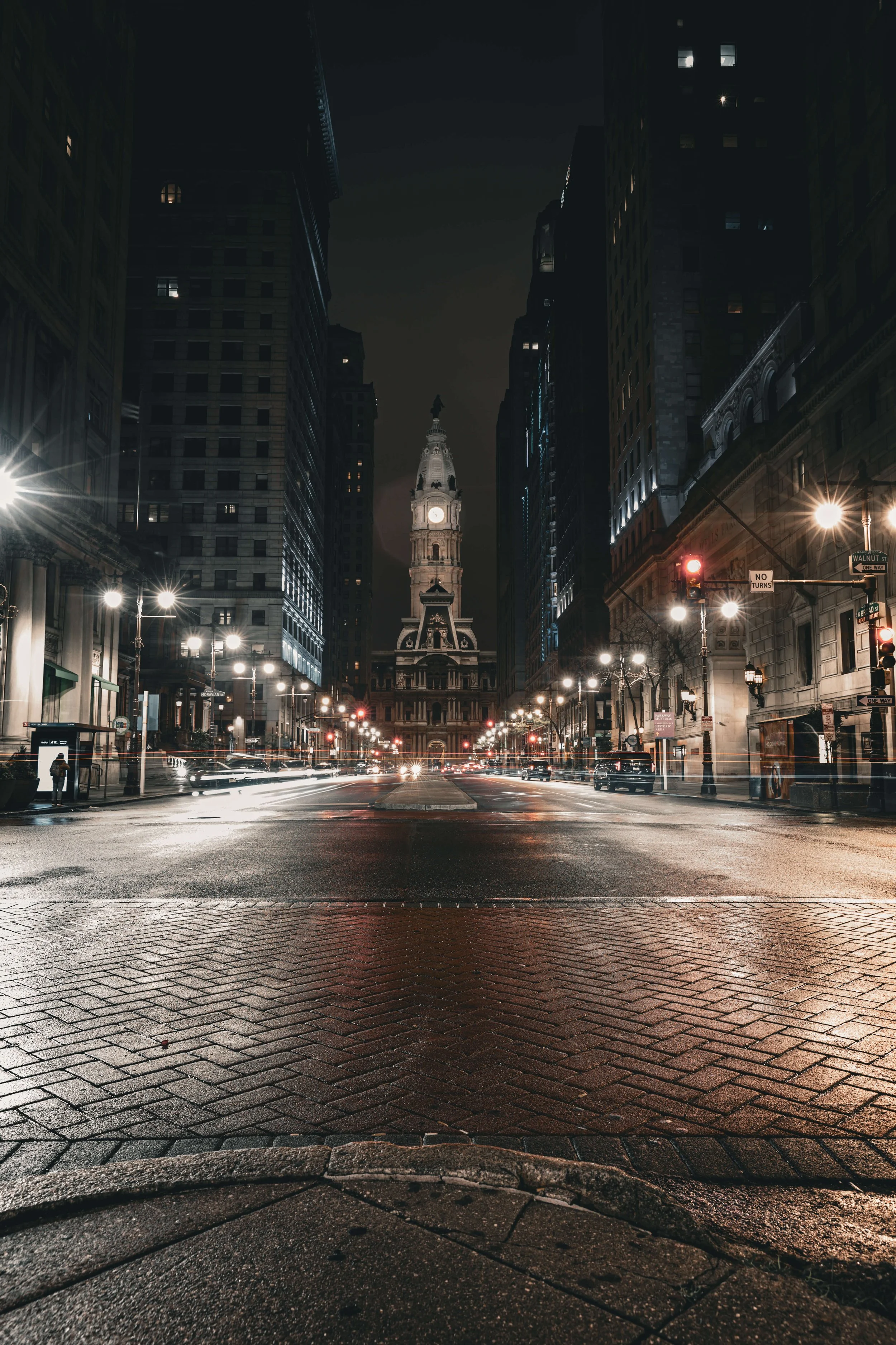 Nighttime city street with historic building featuring a clock tower at the end of the road, surrounded by tall dark buildings, illuminated street lights, and light trails from moving vehicles.