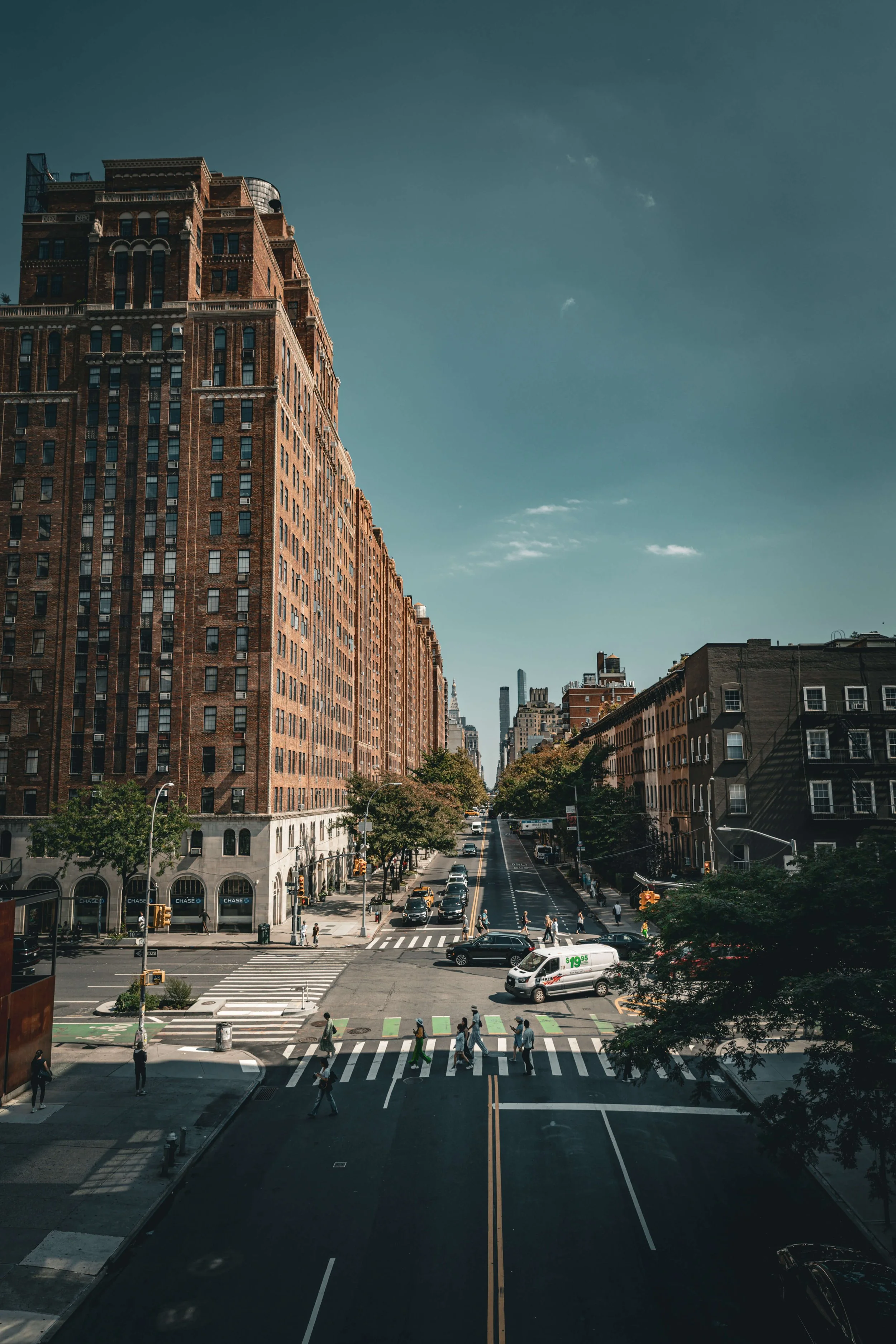 City street with tall brick buildings, cars, and pedestrians crossing the crosswalk on a sunny day.