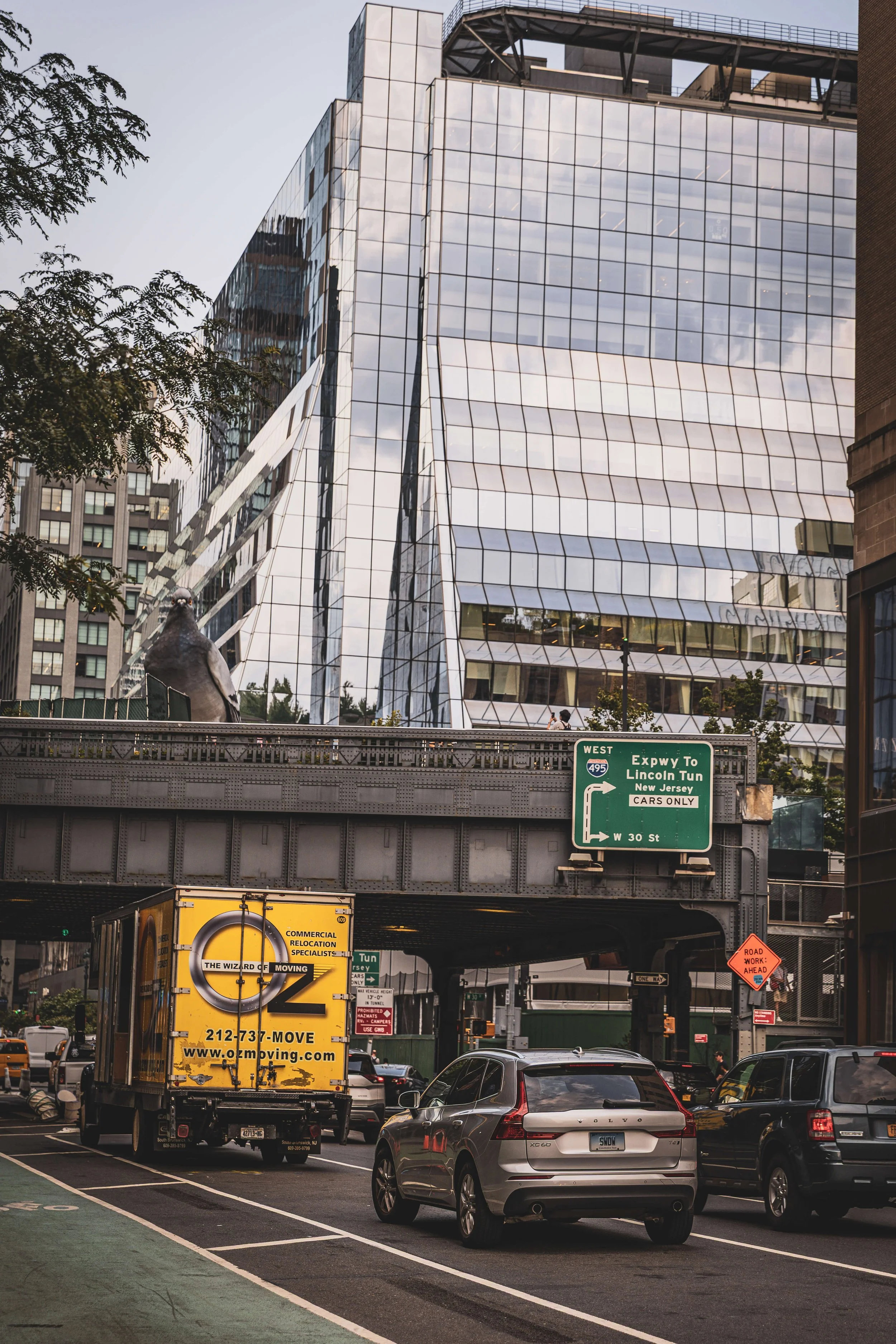 City street scene with cars parked and moving, a large modern glass building with a reflective surface, an overhead highway with green directional signs, a pigeon on the highway barrier, and trees visible in the background.