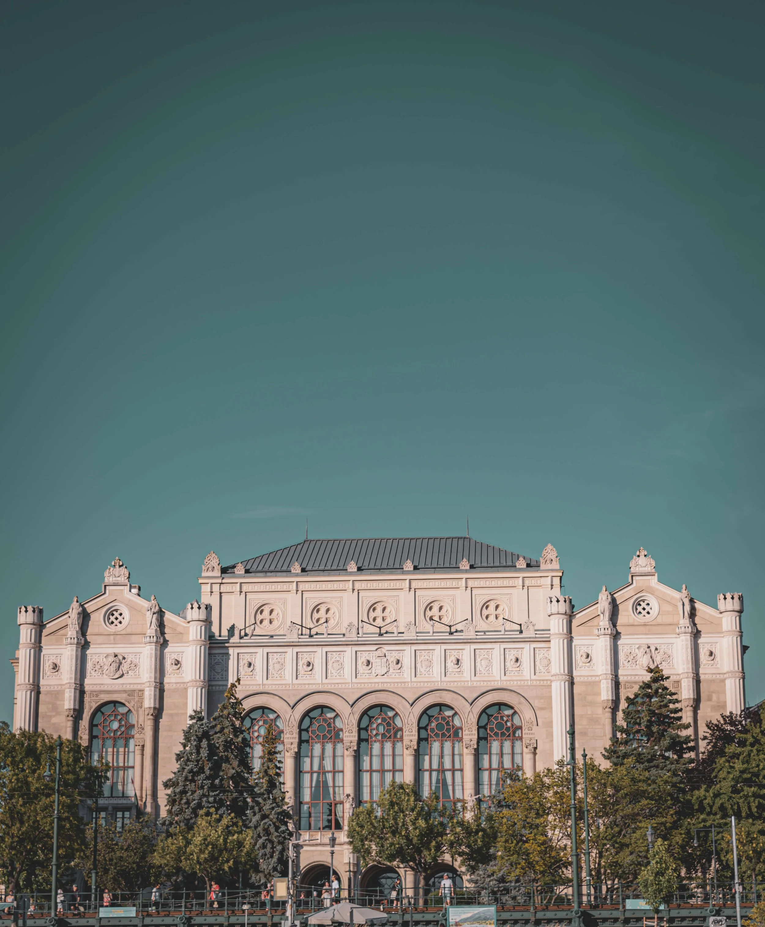 A historic building with intricate architectural details and large arched windows, surrounded by trees and a pedestrian walkway in front.