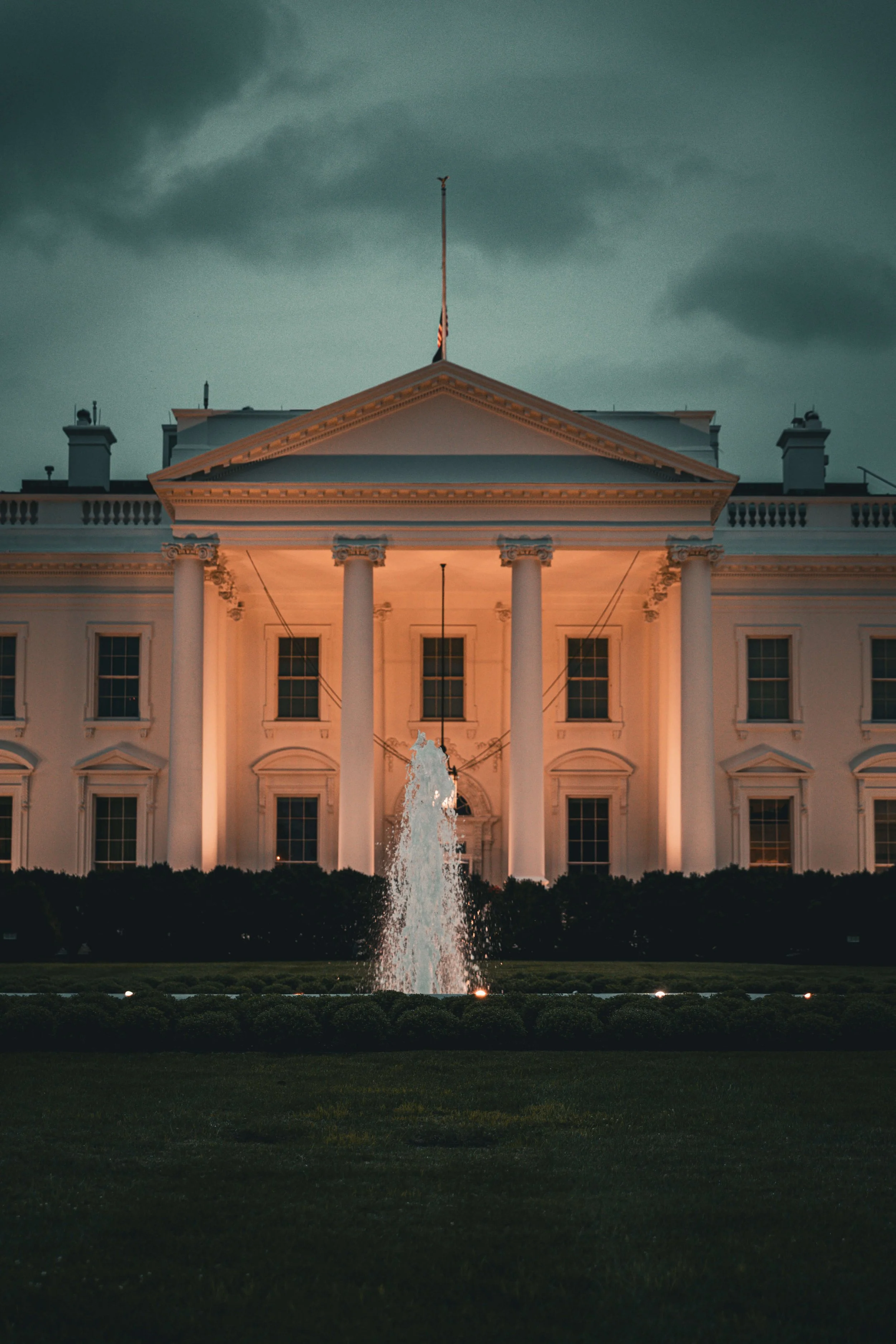 The White House illuminated at dusk with a fountain in front and dark cloudy sky above.