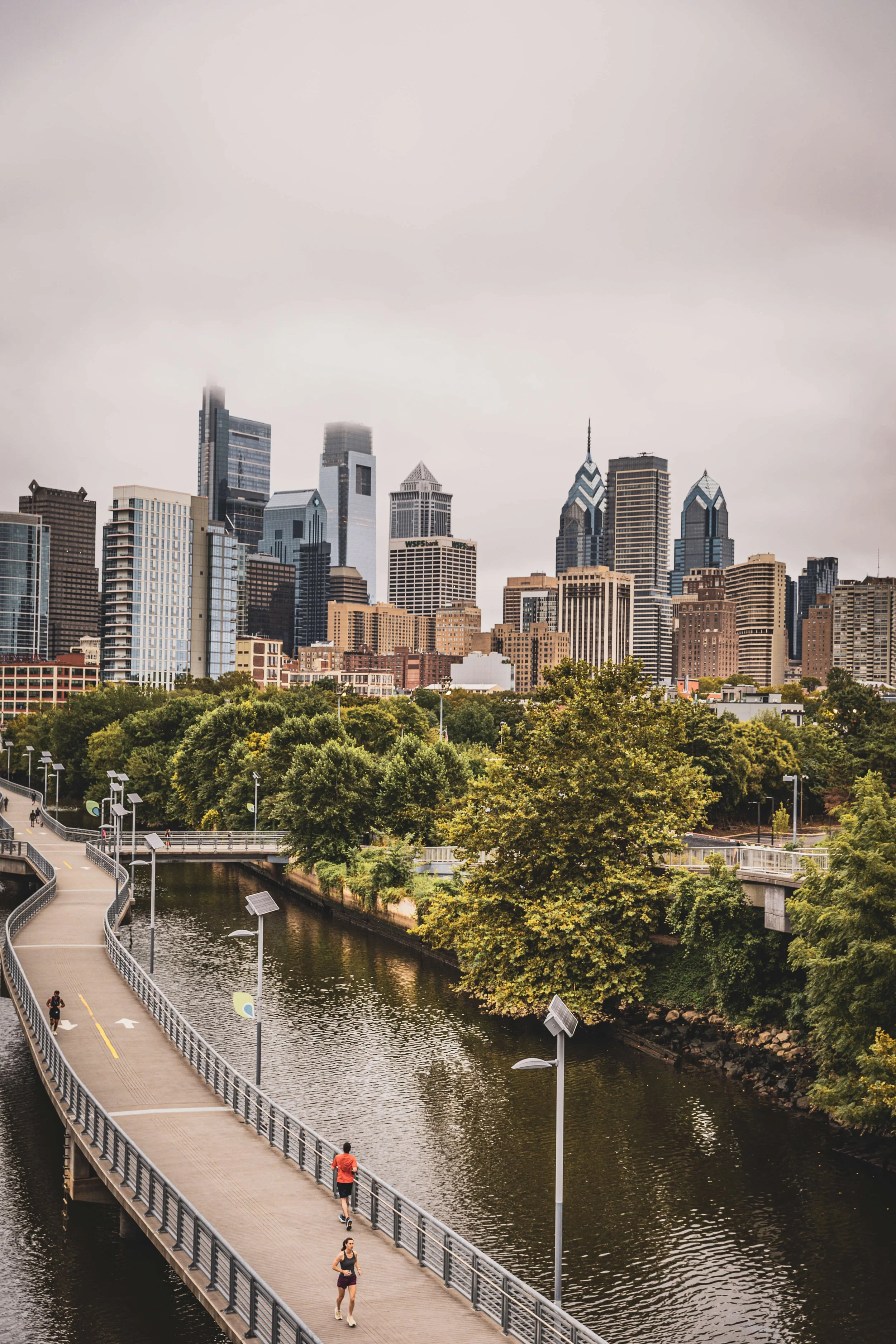 City skyline with tall skyscrapers behind a park with trees and a river, and people running on a bridge.