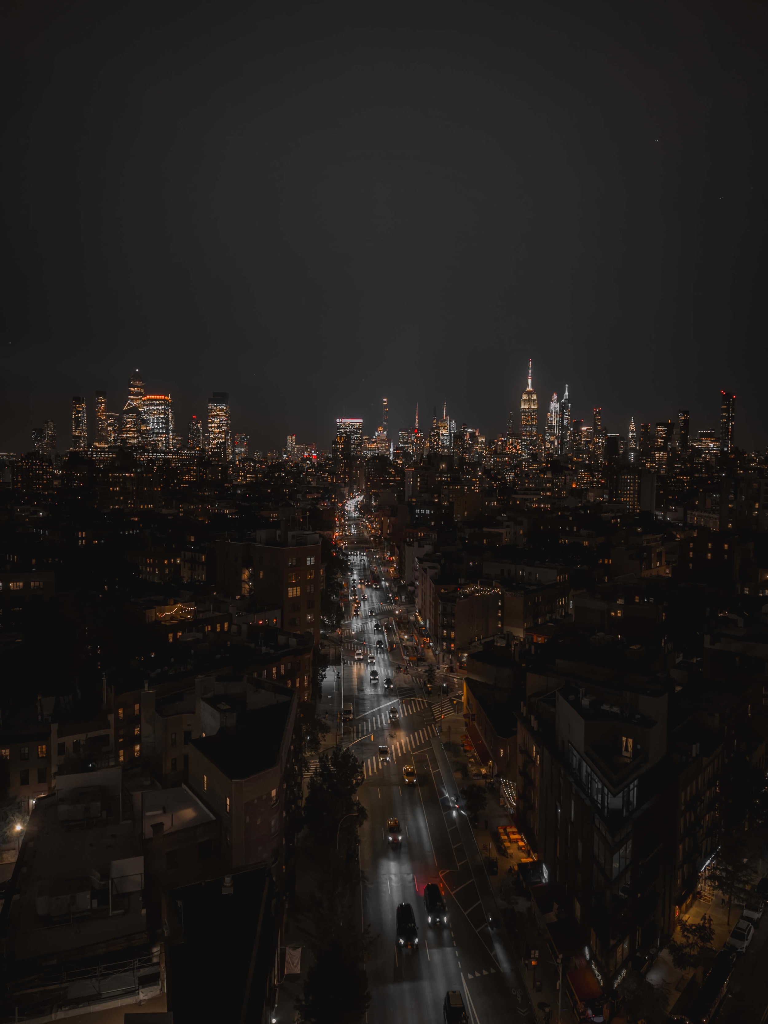 Nighttime cityscape of New York City showing tall illuminated skyscrapers, including the Empire State Building, with busy streets below reflecting city lights.