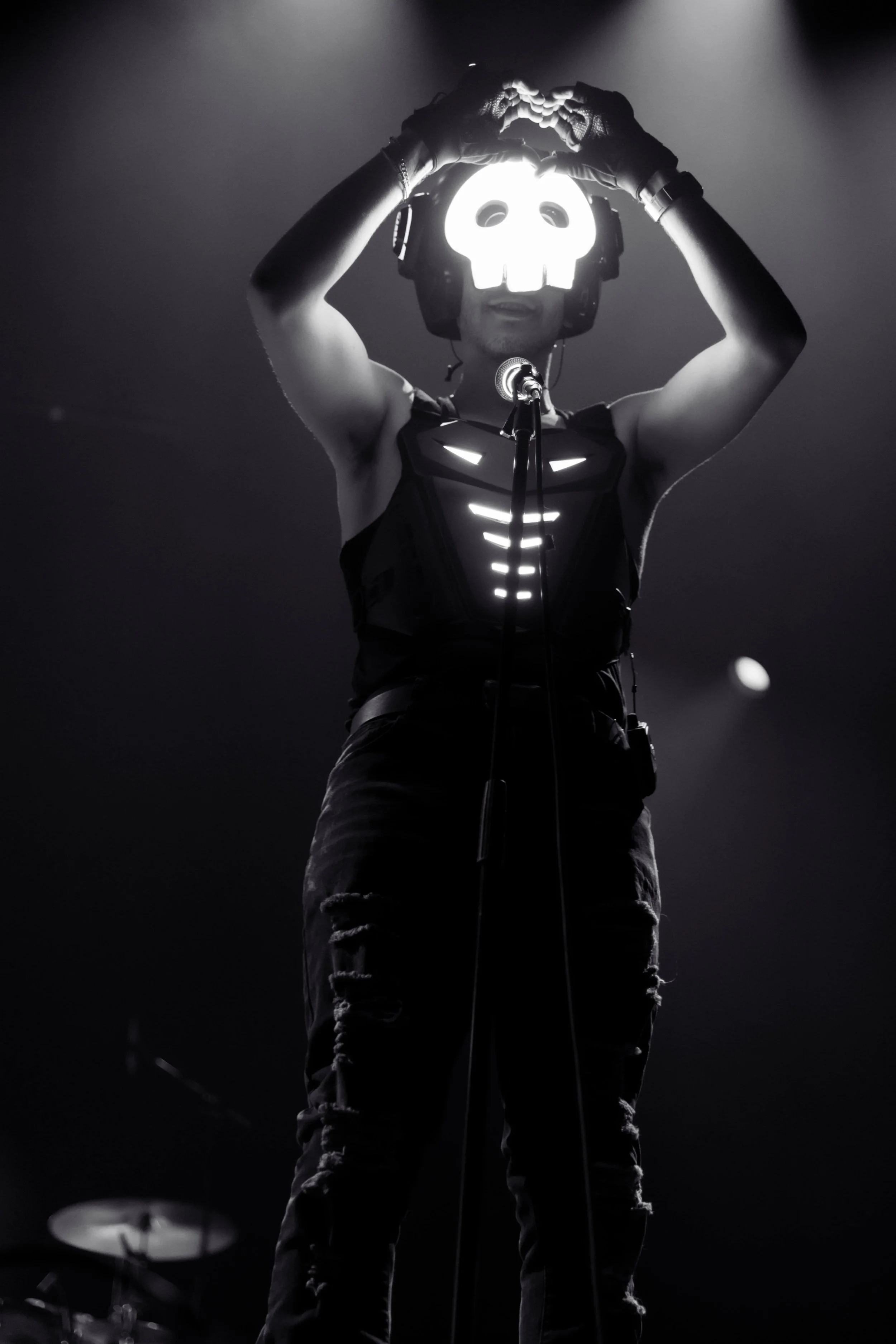 A performer on stage wearing a skull-shaped mask that lights up, a sleeveless shirt with reflective stripes, gloves, and ripped jeans, holding their hands above their head in a heart shape, with a microphone in front of them and faint stage lights in