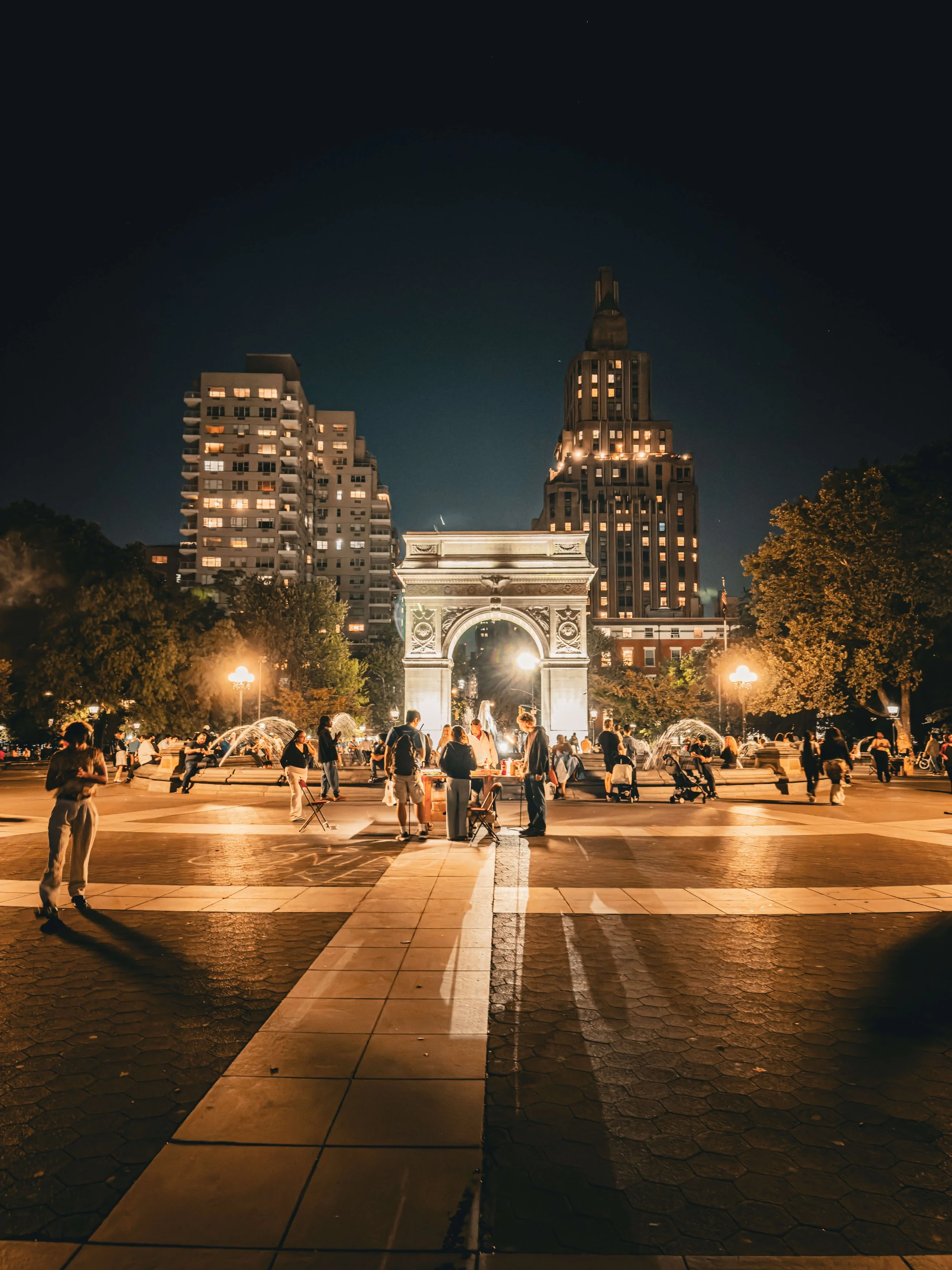 Nighttime scene at a city park with a well-lit arch and fountain, surrounded by tall buildings and people walking or gathering.