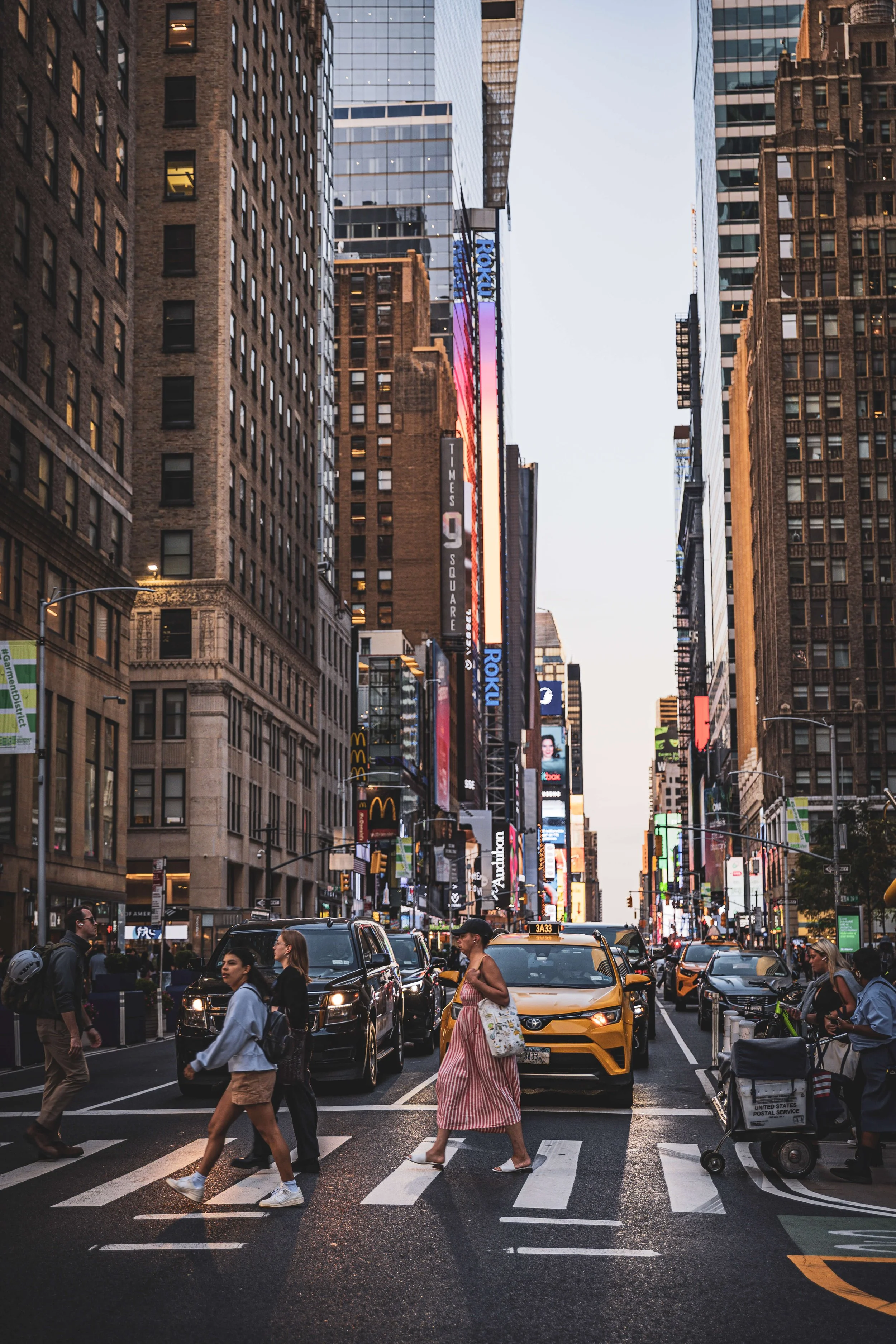 People crossing a busy city street at dusk with tall buildings, illuminated signs, taxis, and other cars.