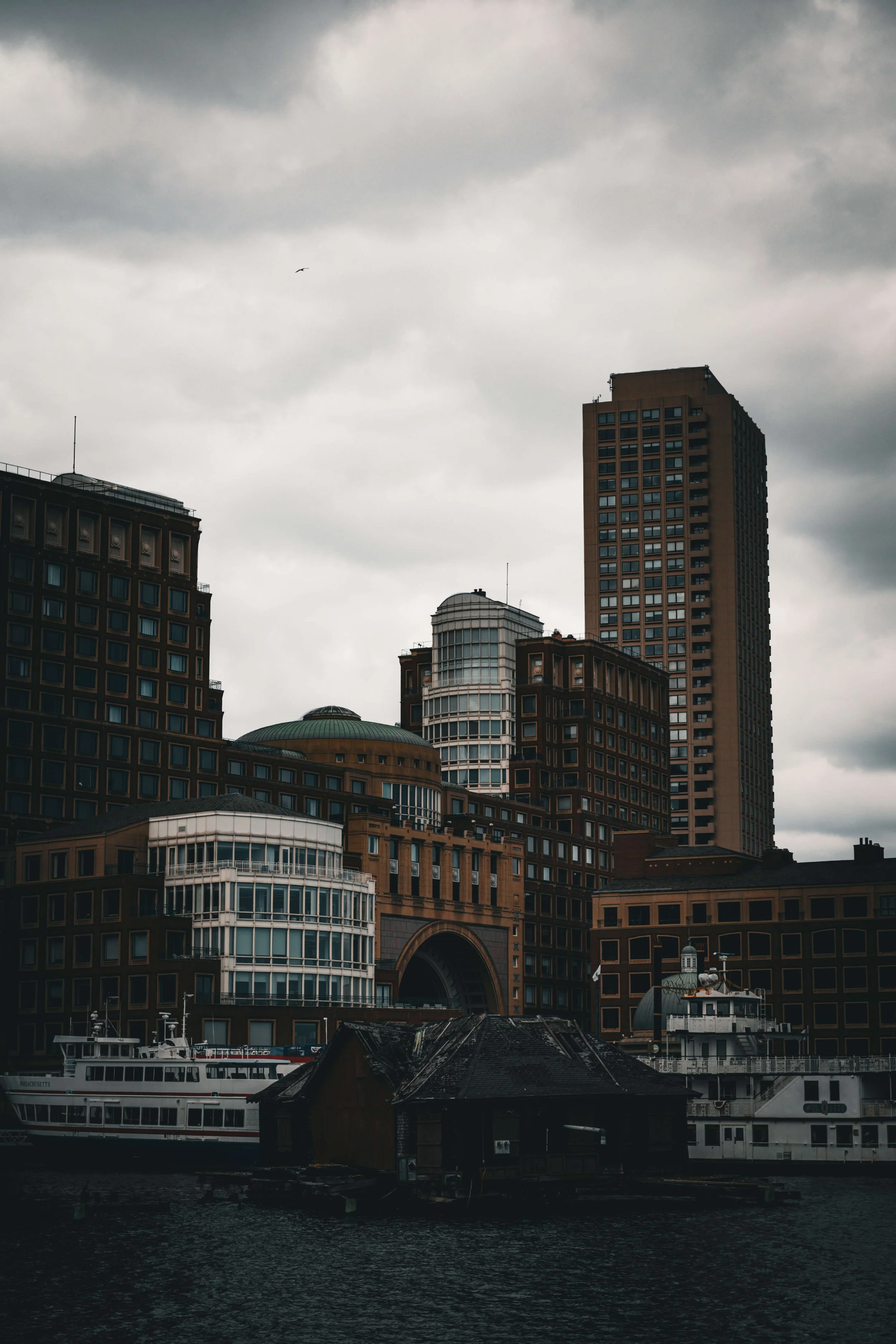 A city skyline under dark, cloudy sky with tall buildings and boats docked on water in foreground.