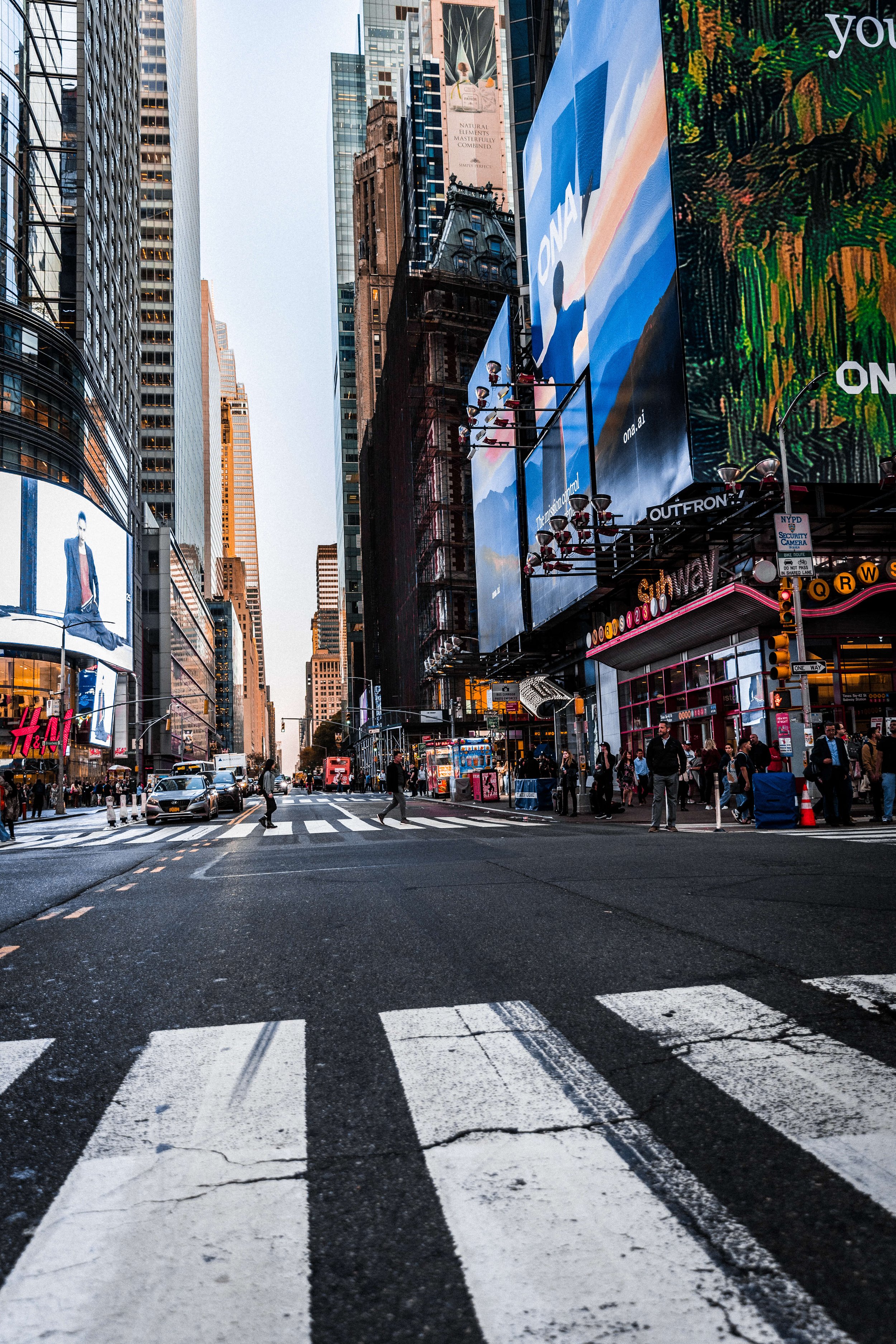 Urban street scene in Times Square, New York City, with pedestrians crossing the crosswalk, tall buildings with billboards, digital screens, and a variety of storefronts and advertisements.