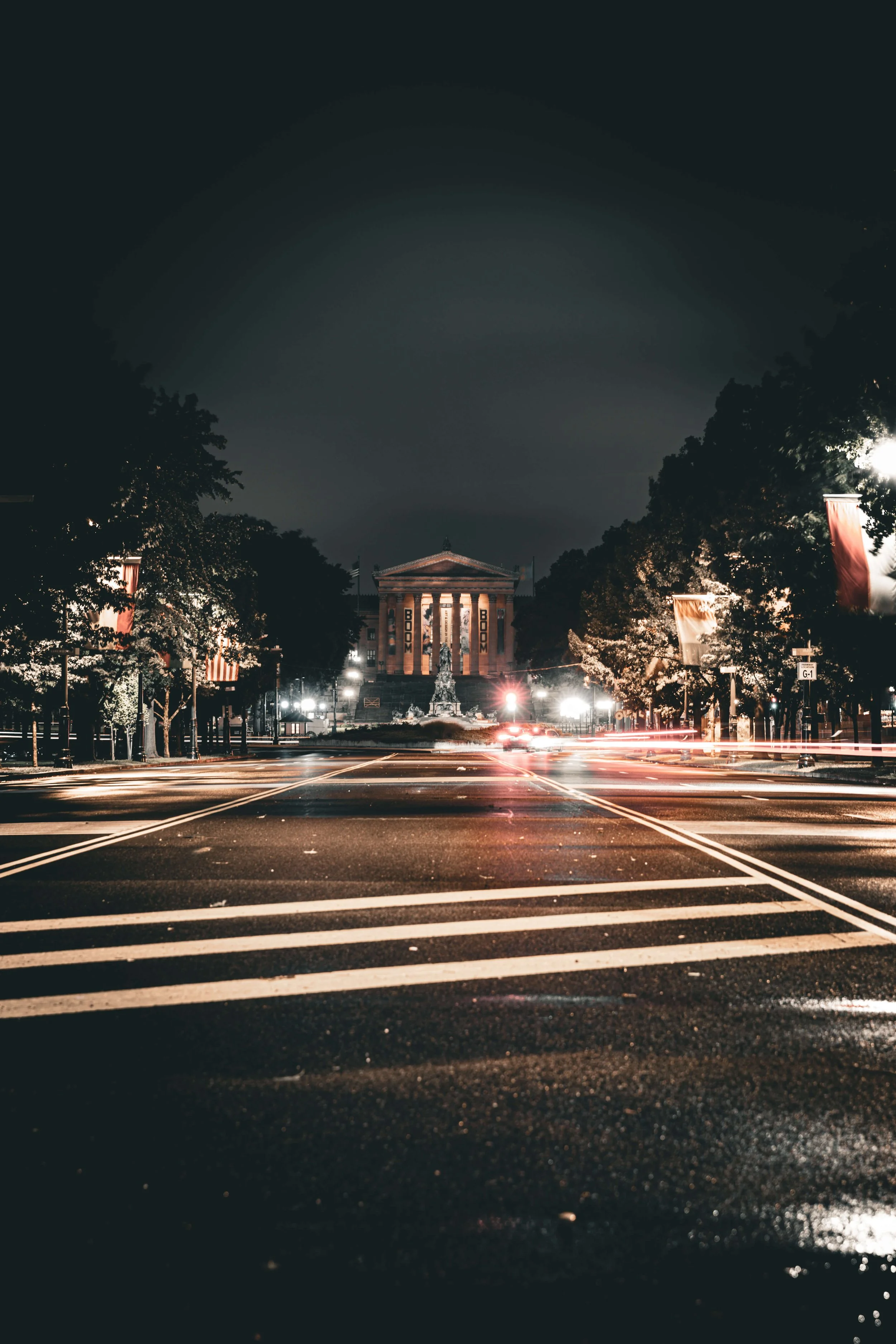 Nighttime view of a city street with empty crosswalk lines, illuminated trees on either side, and a historic building with columns in the background. Lights from vehicles are visible, creating light streaks.