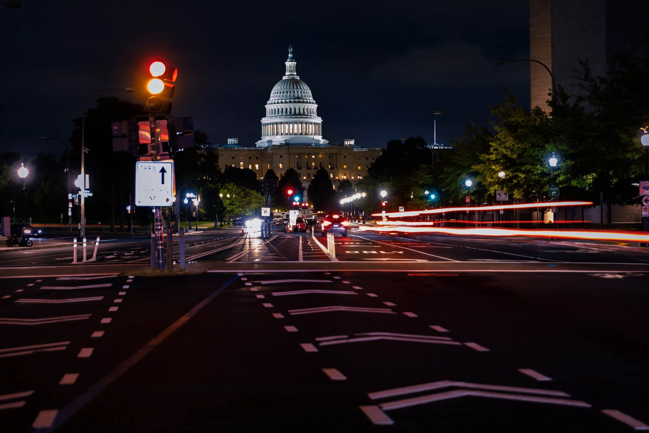 Nighttime view of the U.S. Capitol building in Washington, D.C., with a traffic light showing red and moving car light trails on the street in the foreground.