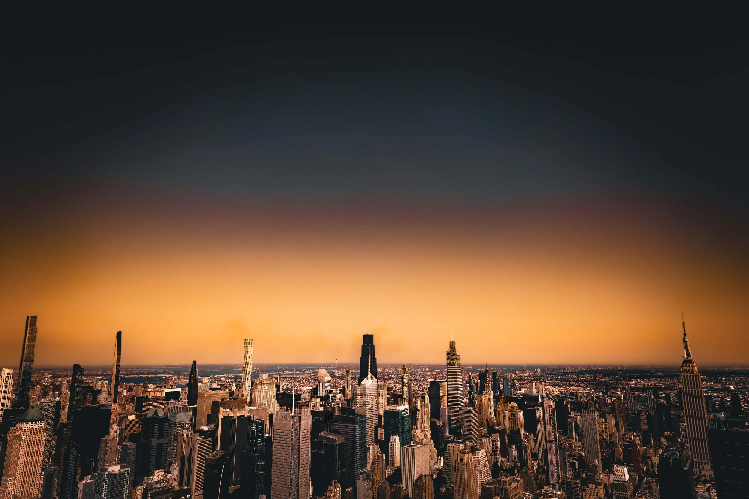 Aerial view of New York City skyline at sunset with a gradient sky transitioning from dark blue to orange, featuring iconic skyscrapers including the Empire State Building.