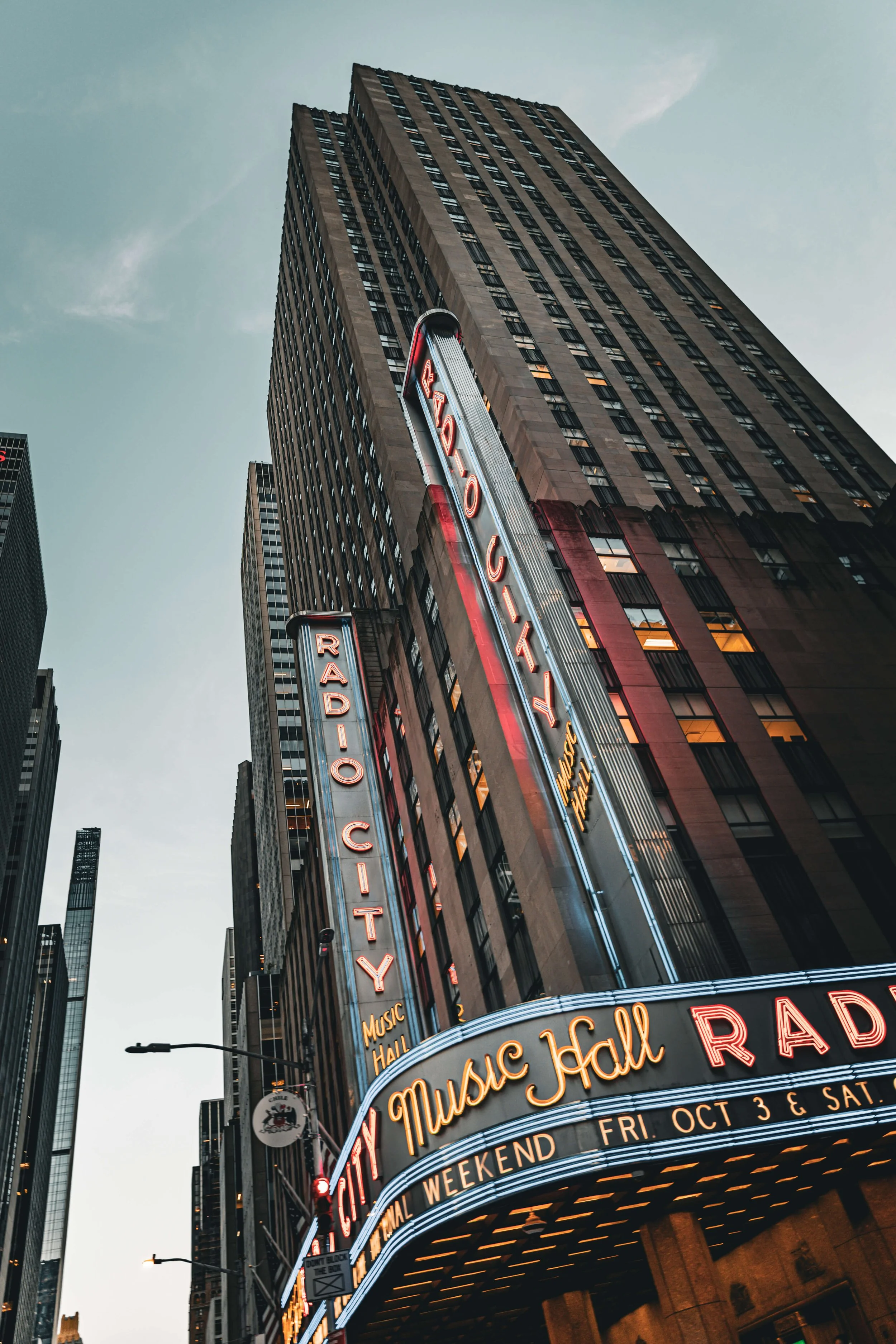 Vertical shot of the Radio City Music Hall in New York City, with illuminated marquee signs and tall skyscrapers in the background.