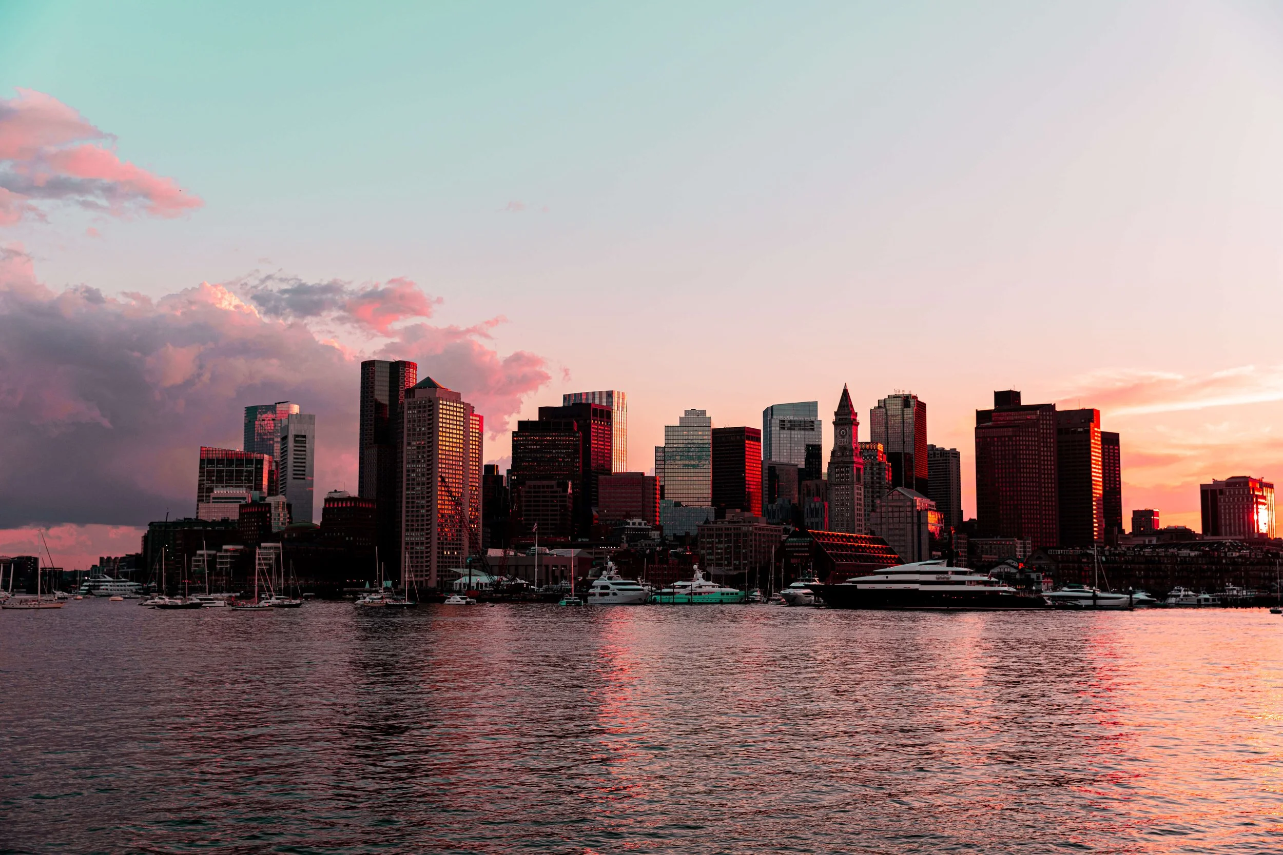 Sunset over a city skyline with high-rise buildings reflected in the water, boats docked at a marina in the foreground.