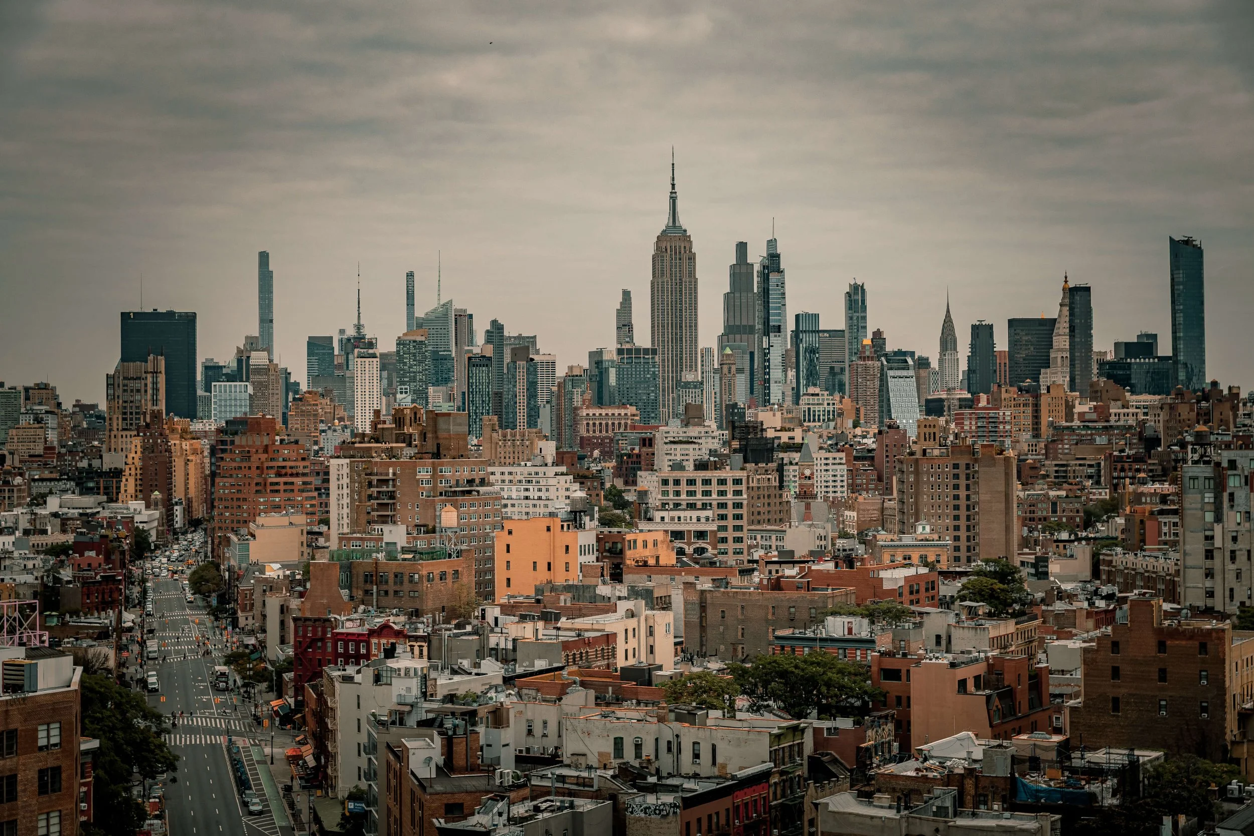 City skyline of Manhattan, New York, with tall skyscrapers including the Empire State Building under a cloudy sky.