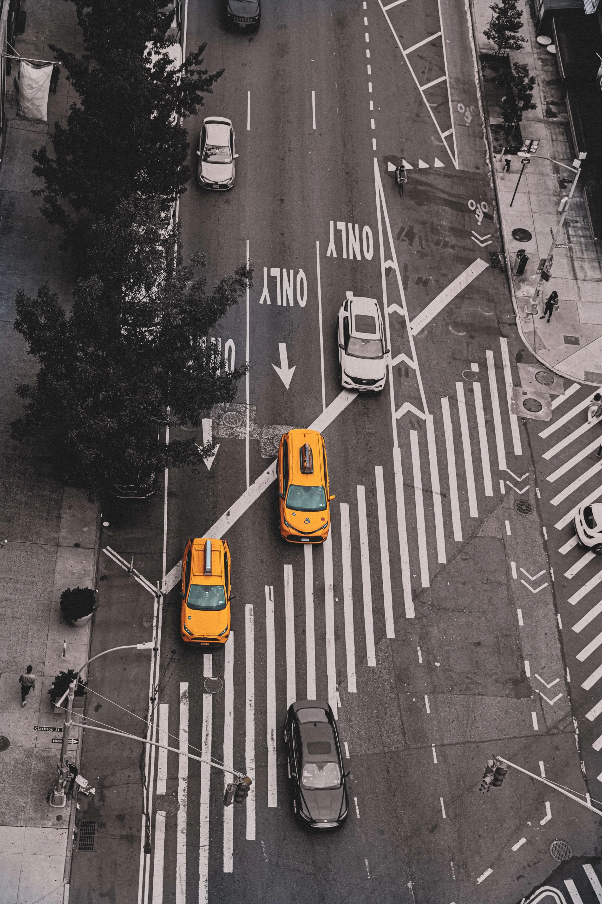 Aerial view of a city street with cars, including two yellow taxis, parked along the sidewalk and moving on the road. Road markings indicate lane directions, and there are crosswalks, bike lanes, and a few pedestrians on the sidewalk.