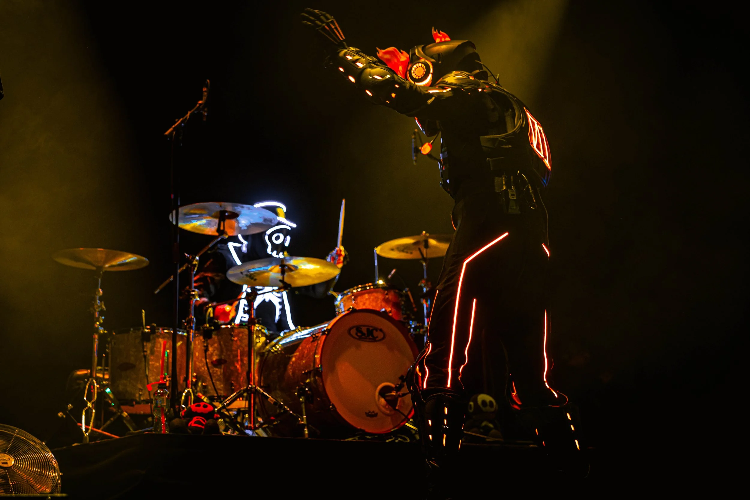 A performer dressed in a futuristic, LED-lit costume with fiery flame accents, performing with a drum set on a dark stage, illuminated by colorful lights.